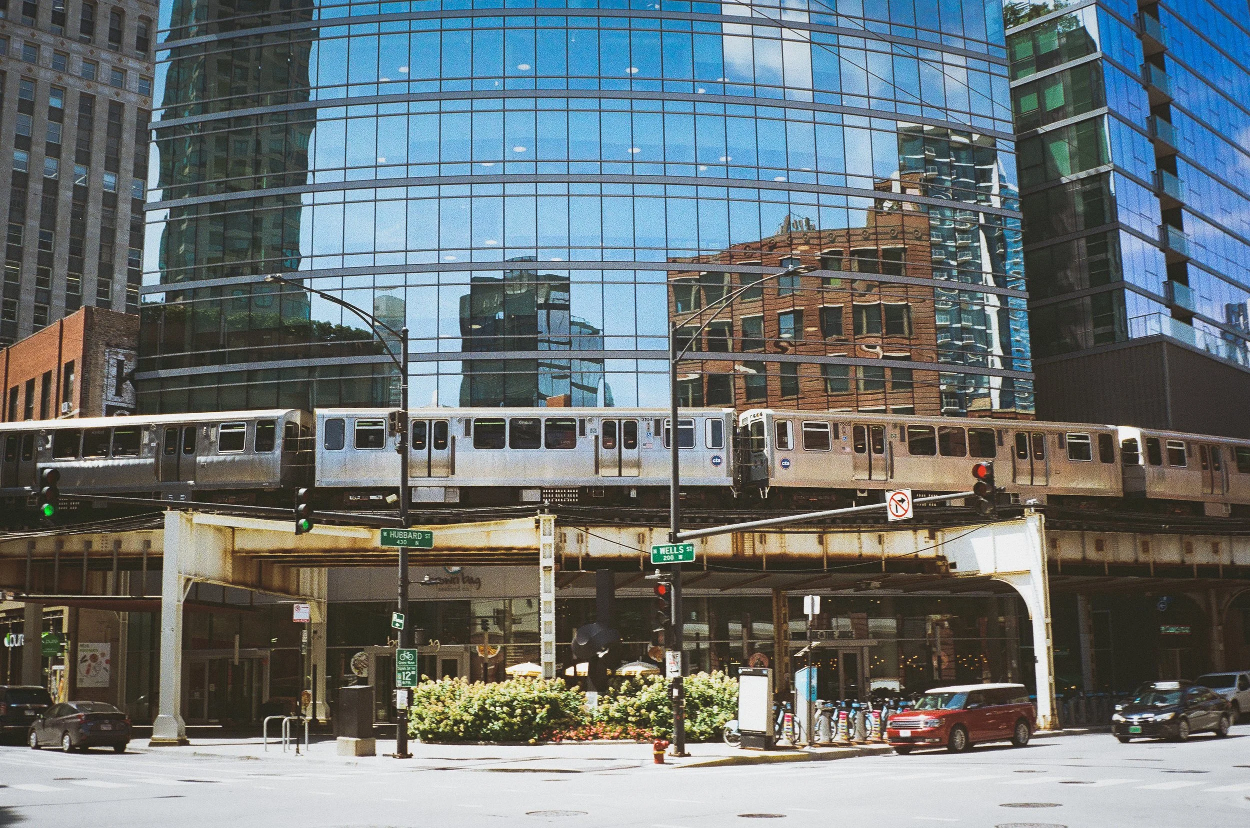 A city street scene with a train above ground and modern glass buildings reflected on the windows behind it. Cars and pedestrians are visible below, along with street signs and a bicycle rack.