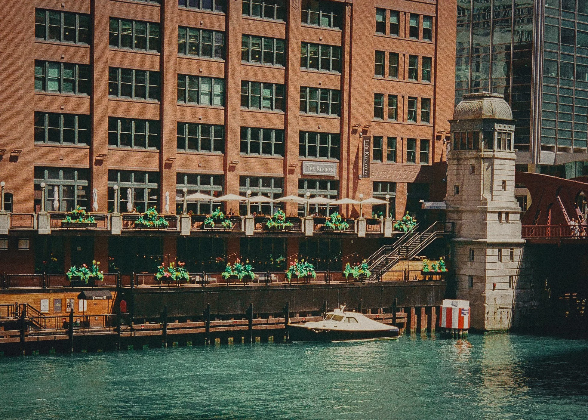 Waterfront building with brick facade, outdoor dining area with umbrellas, stone tower with stairs, docked boat, and a waterway.