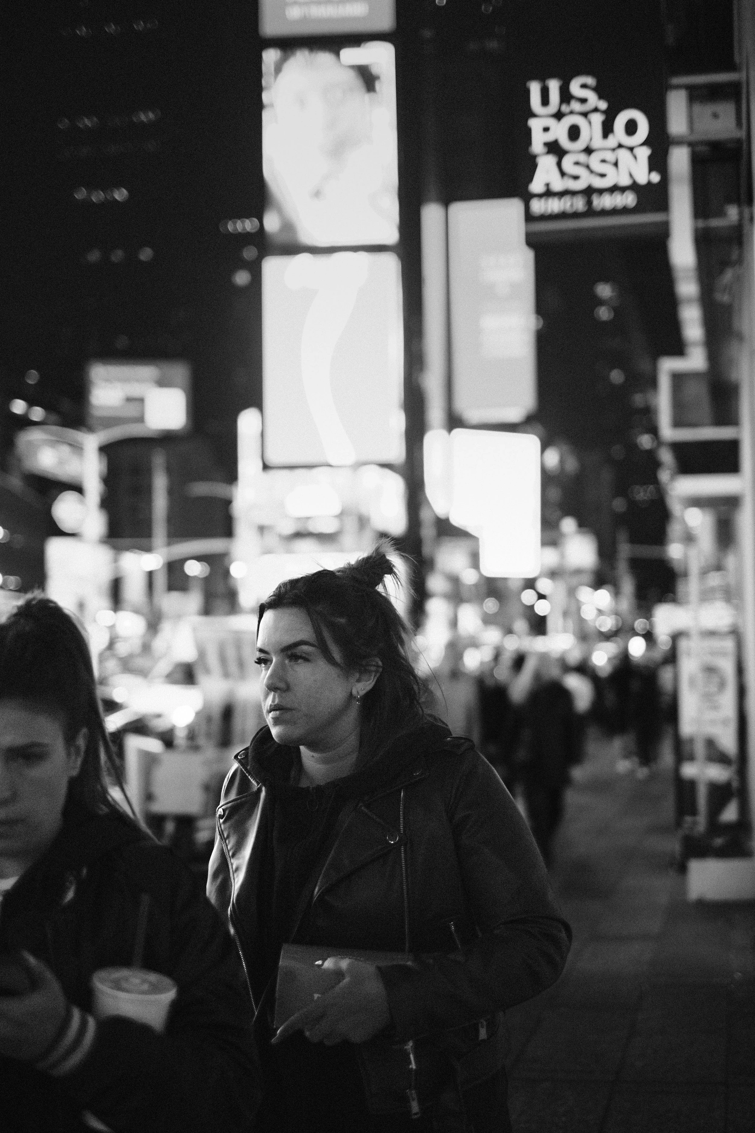 A black and white photo of a woman with dark hair in a messy bun, wearing a leather jacket, walking on a city street at night. Bright city lights and illuminated signs are visible in the background, including a sign for U.S. Polo Assn.