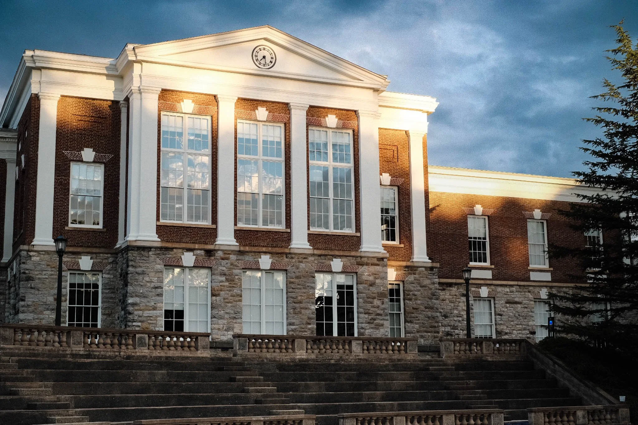 A large brick and stone building with white trim, multiple windows, classical columns, steps, and street lamps in front, under a partly cloudy sky.