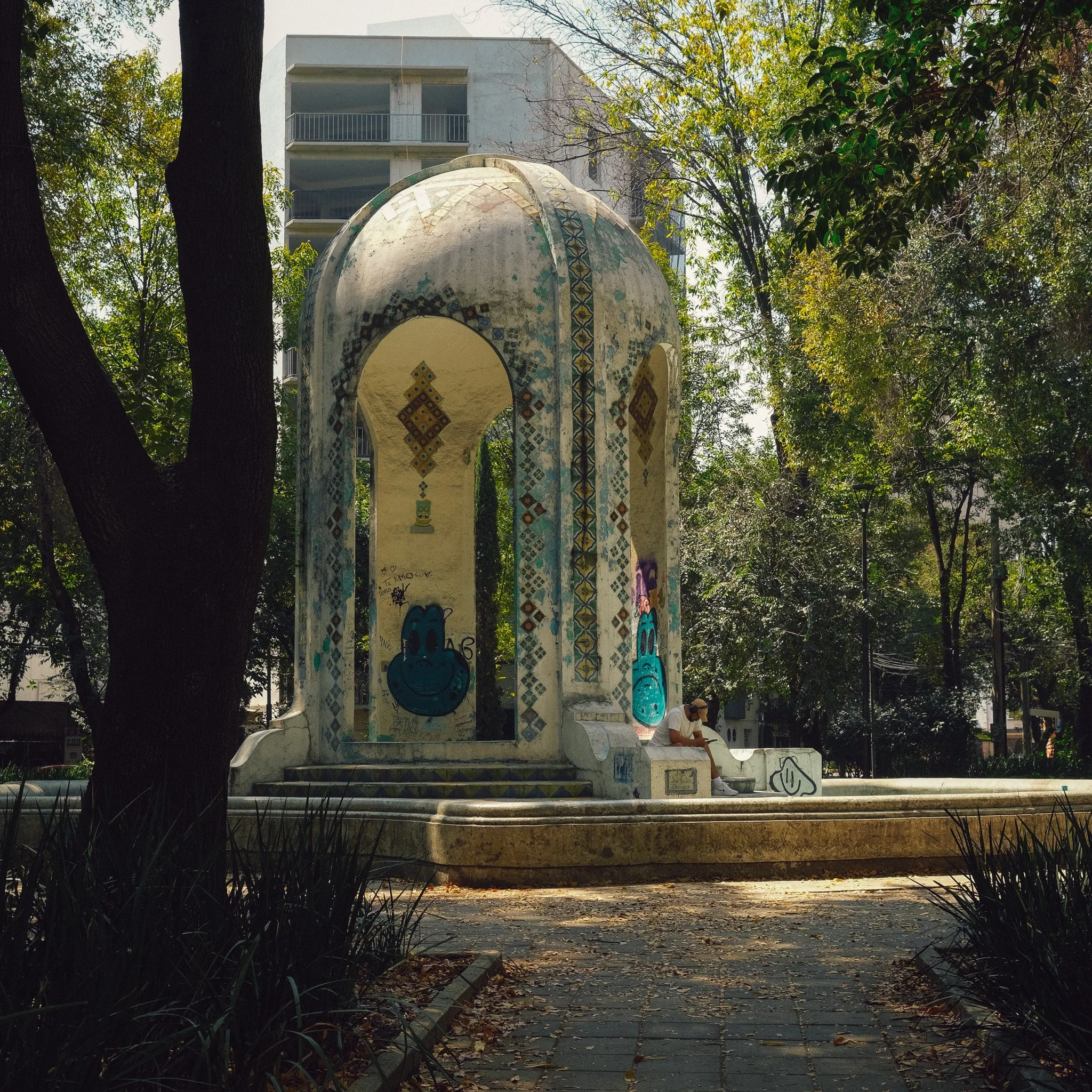 A decorative tiled pavilion in a park with graffiti art, surrounded by trees, and a person sitting on the pavilion steps wearing a white shirt and a hat.