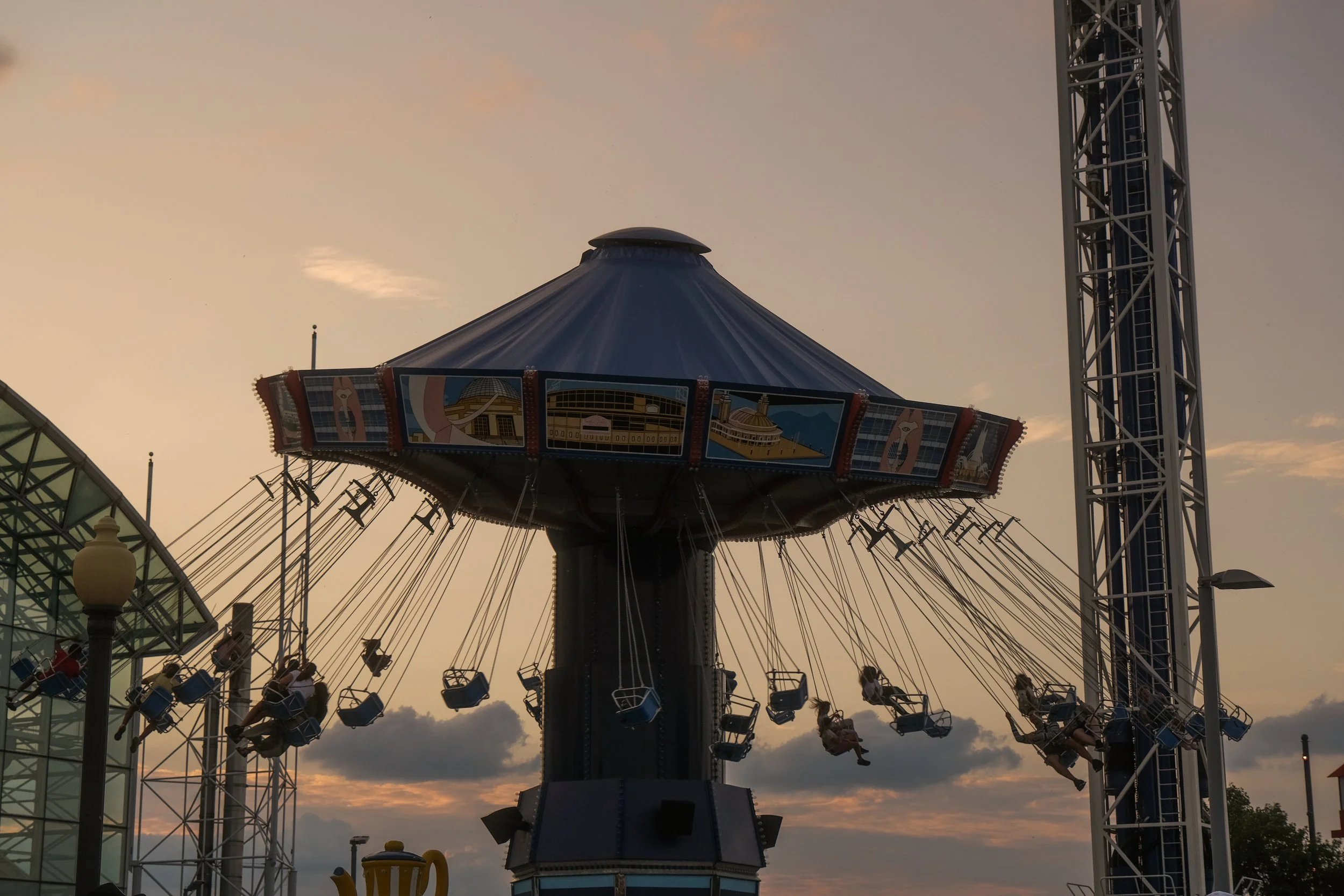 An amusement park swing ride at sunset with children riding the swings, a large blue tent-like structure on top, and a metal tower on the right side.