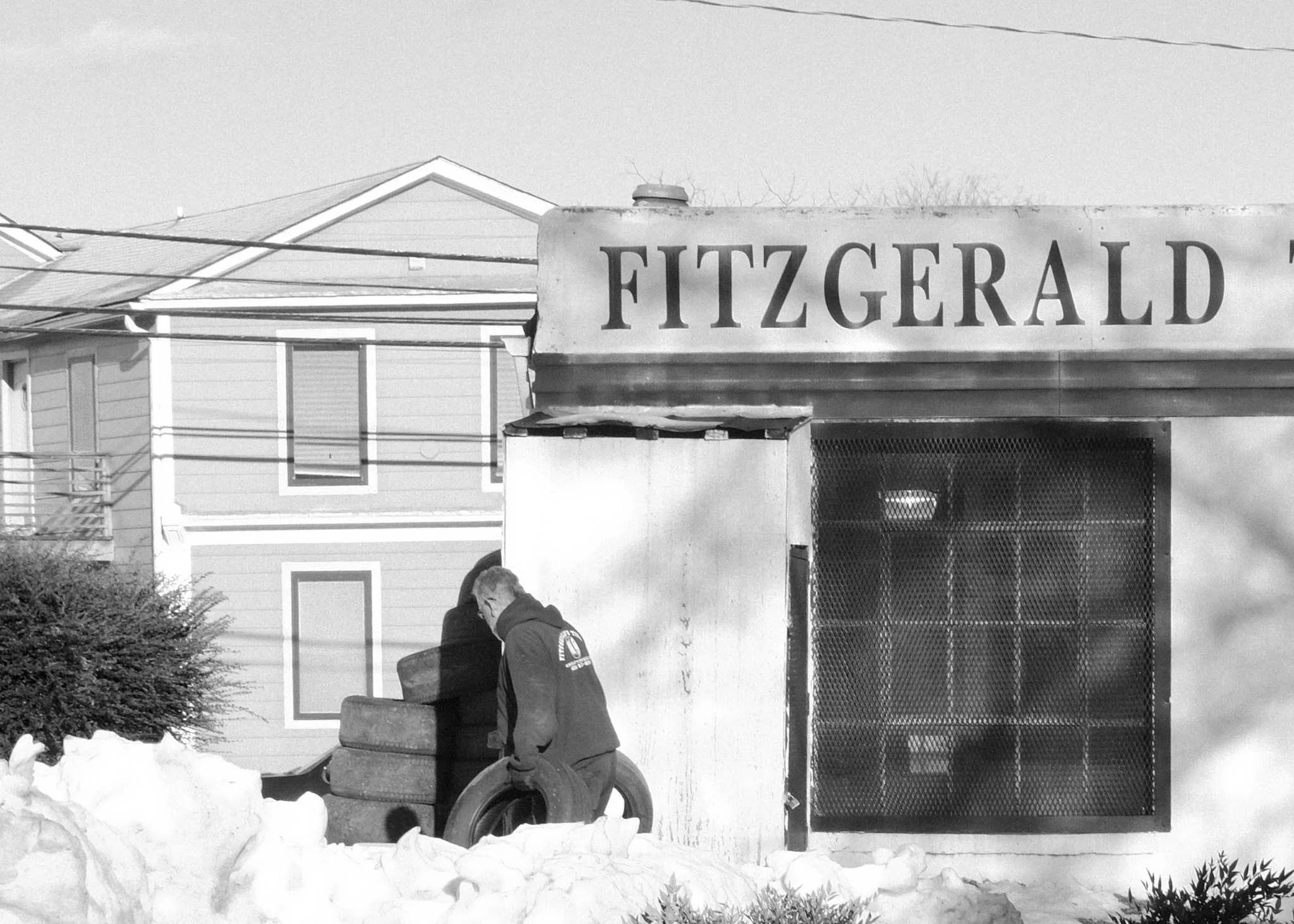 A man in a hoodie stands outside a small, closed business with a sign that reads 'Fitzgerald.' The building has a large window with metal bars, and snow is piled up around the area.