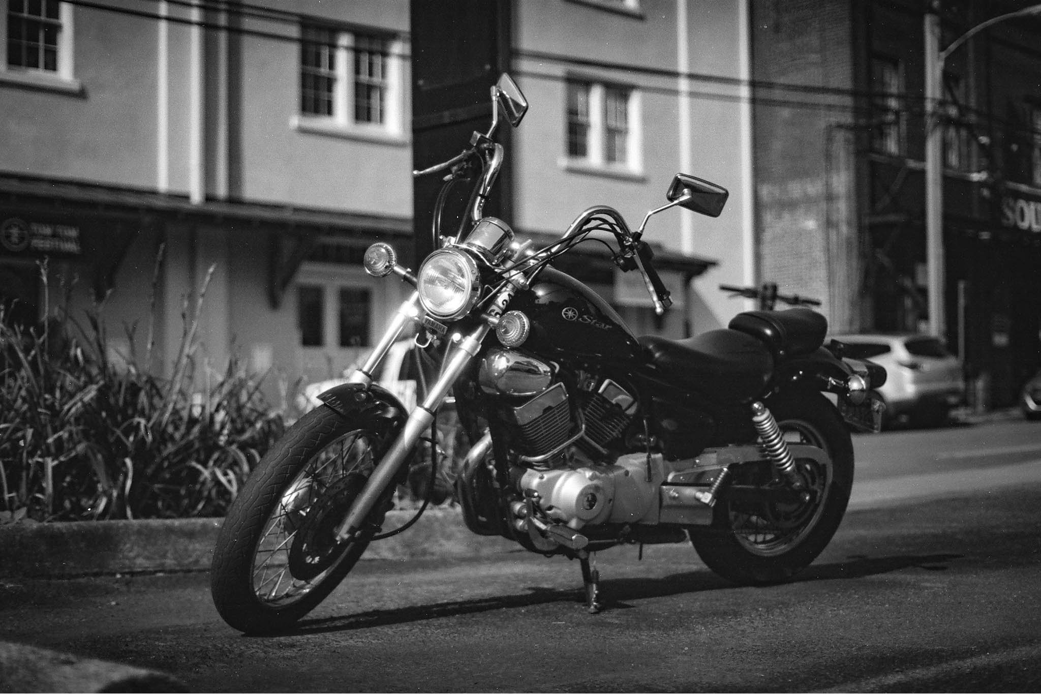 A black and white photo of a cruiser motorcycle parked on the street at night, with apartment buildings and parked cars in the background.