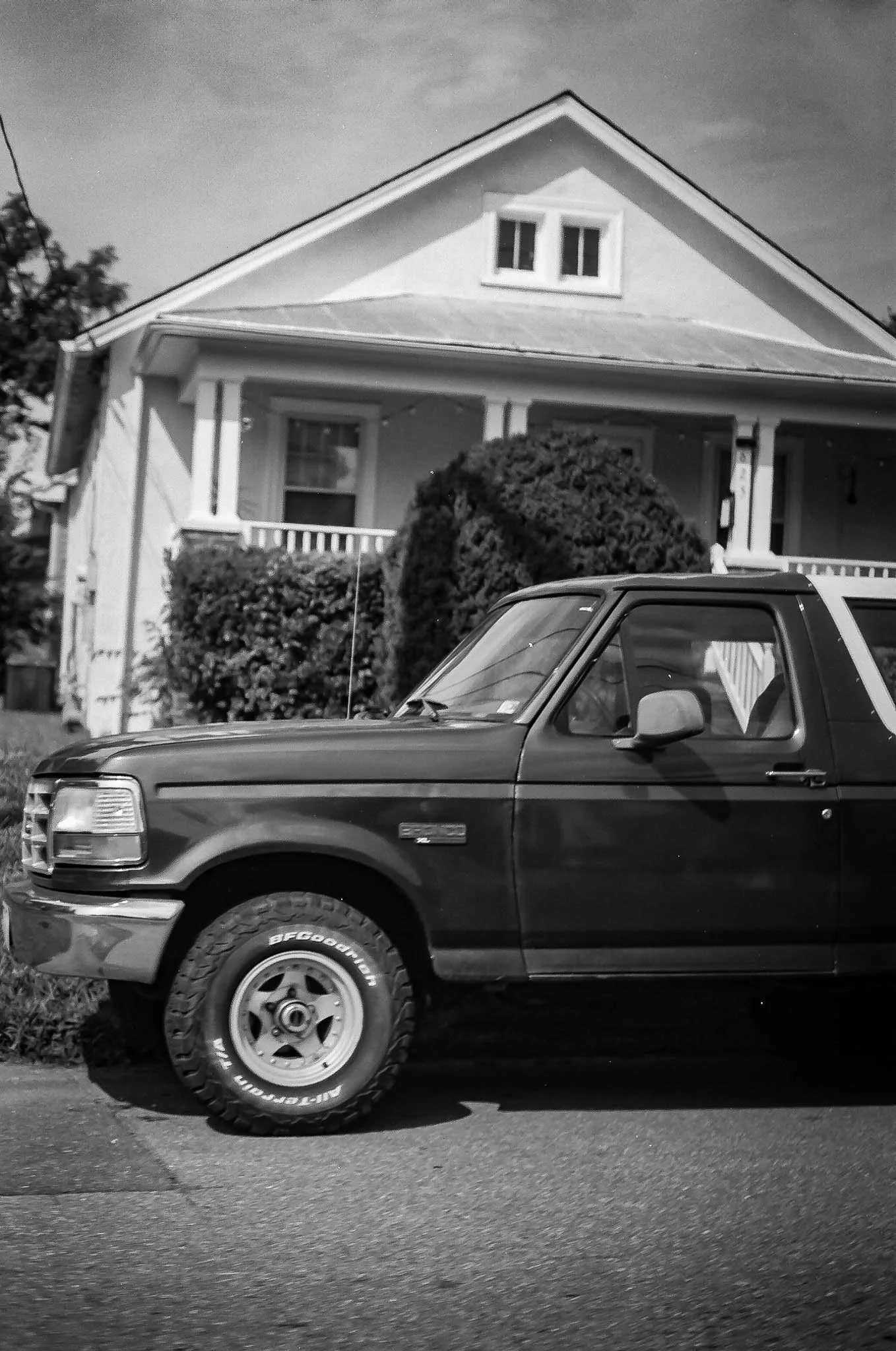 A black pickup truck parked in front of a residential house with a porch and shrubbery in a black and white photo.