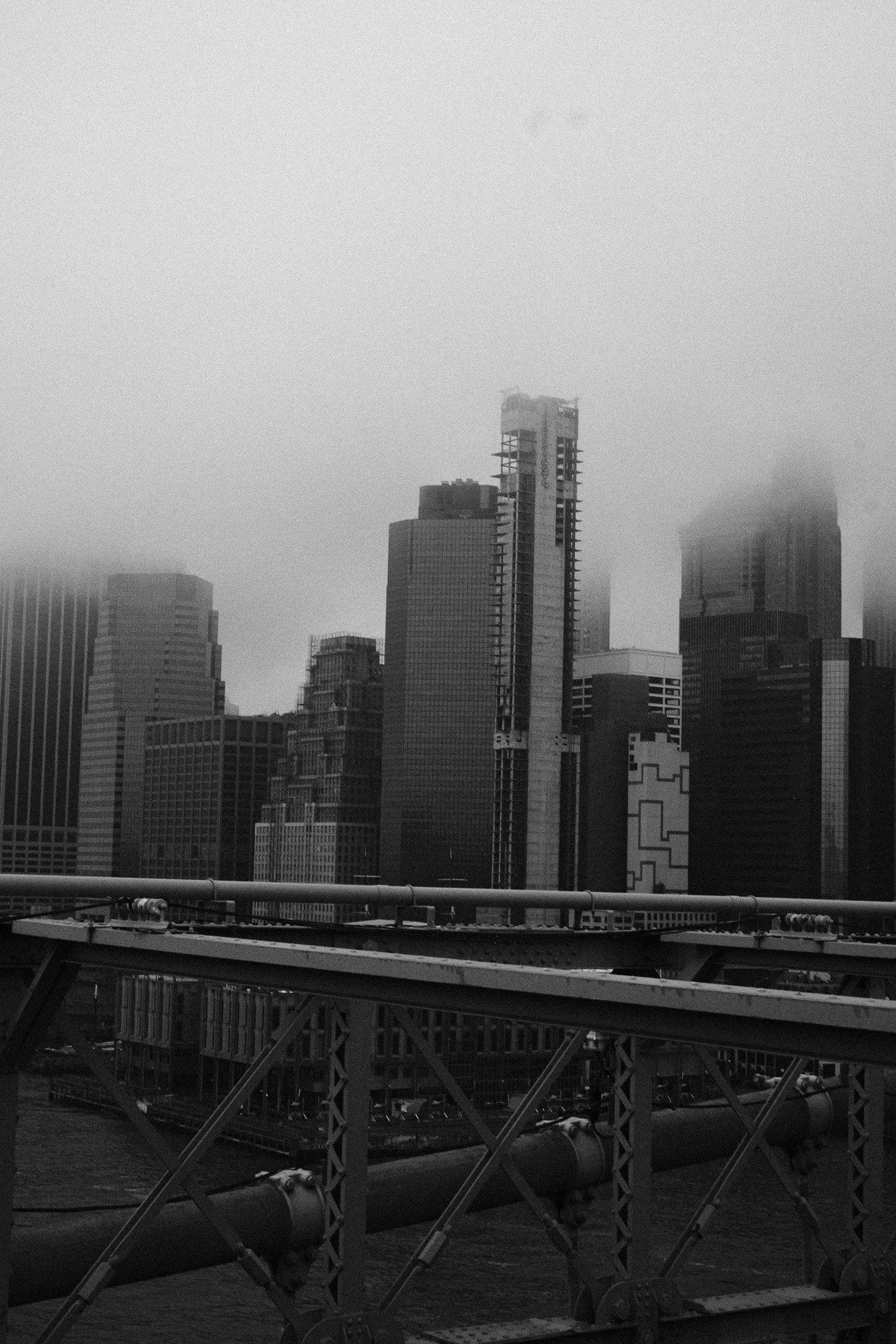 Black and white photo of a city skyline with tall buildings, some partially obscured by fog, and a bridge in the foreground.