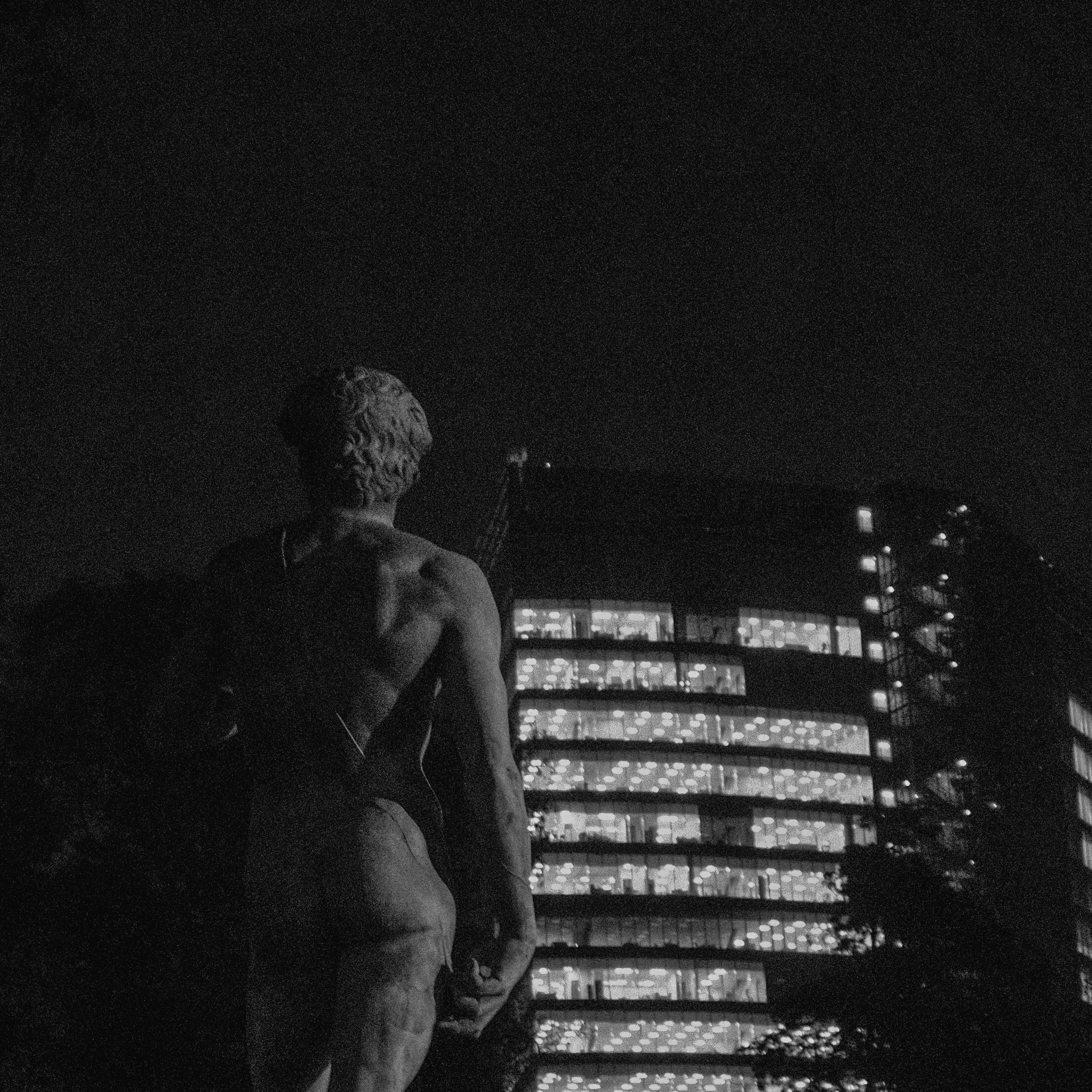 A black and white photo of a classical statue of a man with curly hair, facing away from the camera, with a modern, lit office building in the background at night.