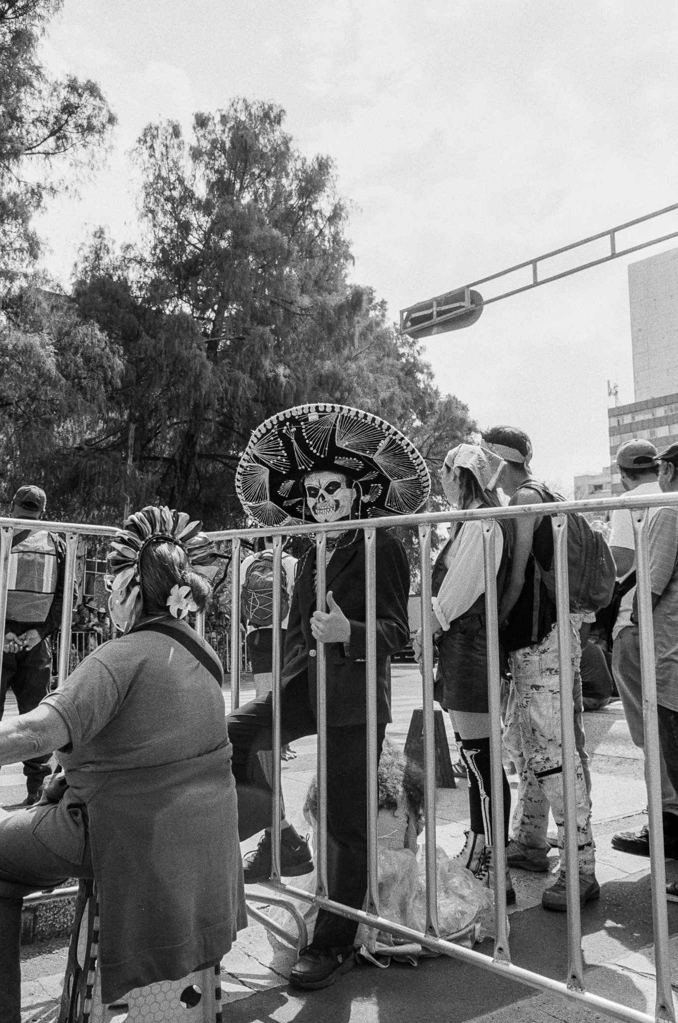 A person in a skeleton costume wearing a sombrero stands behind a barricade during a parade, with other people and trees in the background.