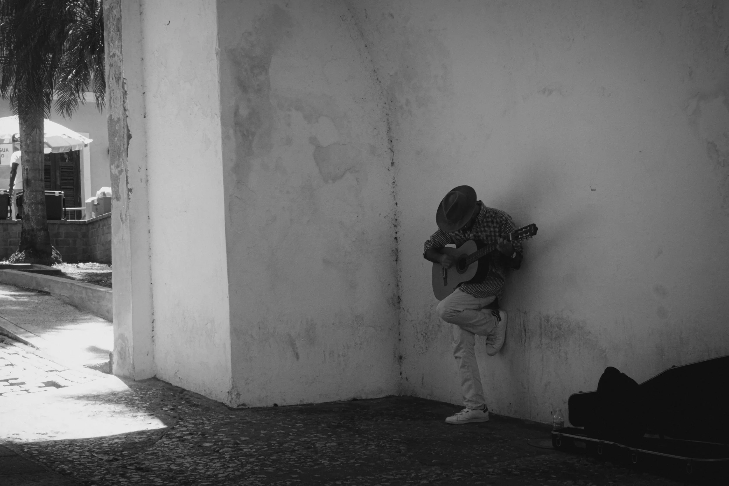 A street musician wearing a hat and casual clothes playing an acoustic guitar against a weathered wall, with a closed guitar case on the ground nearby and some water bottles placed next to it.