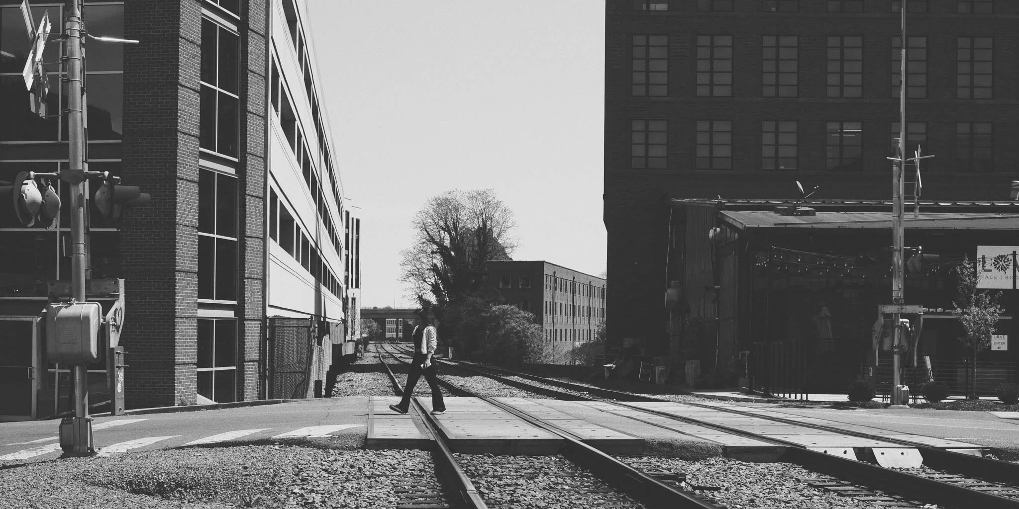 A man in formal attire, walking across railroad tracks in an urban area, with tall buildings on either side and a tree in the background.