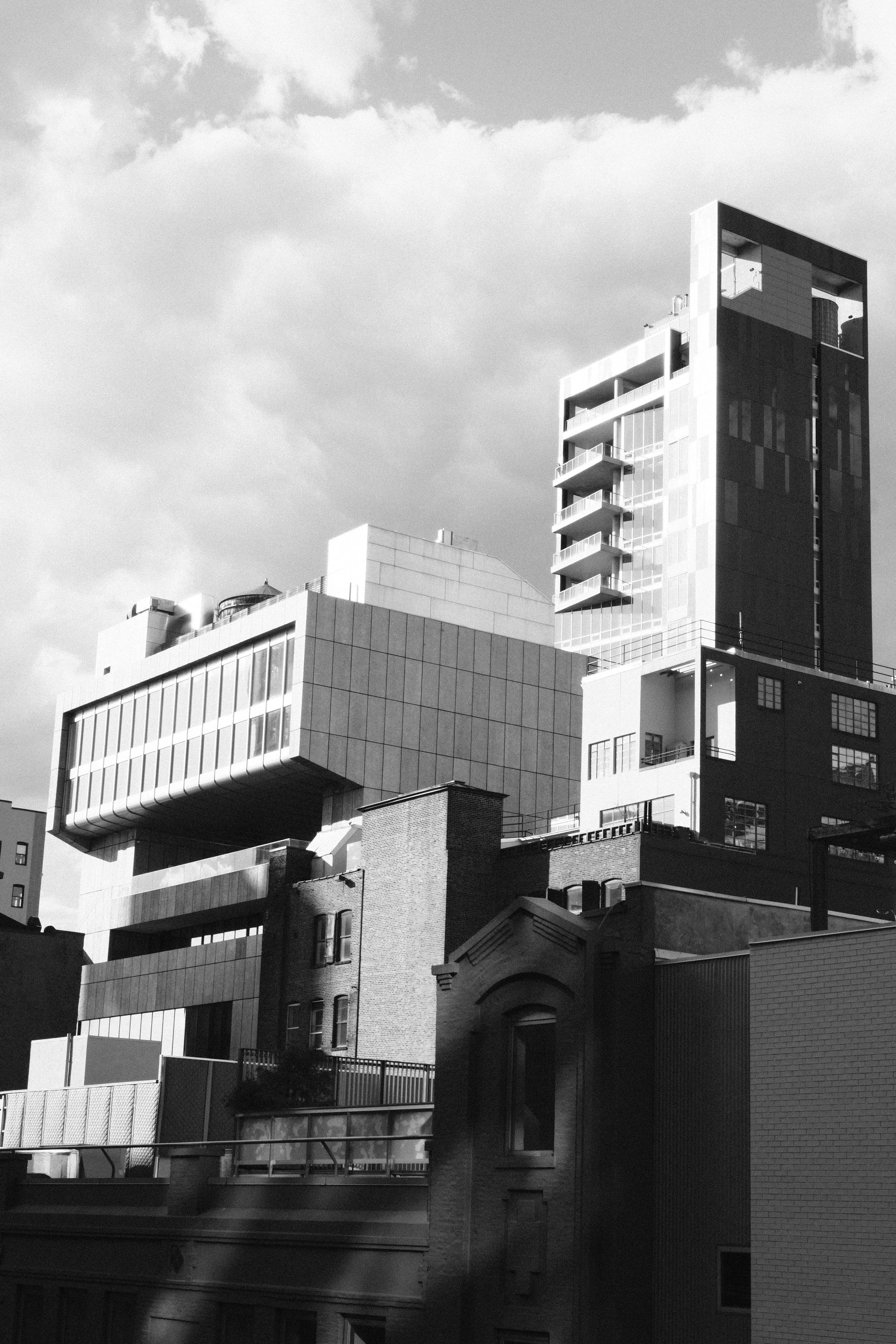 Black and white photo of modern city buildings with a mix of old and new architecture, including a tall high-rise with balconies and a unique cantilevered structure, with cloudy sky overhead.