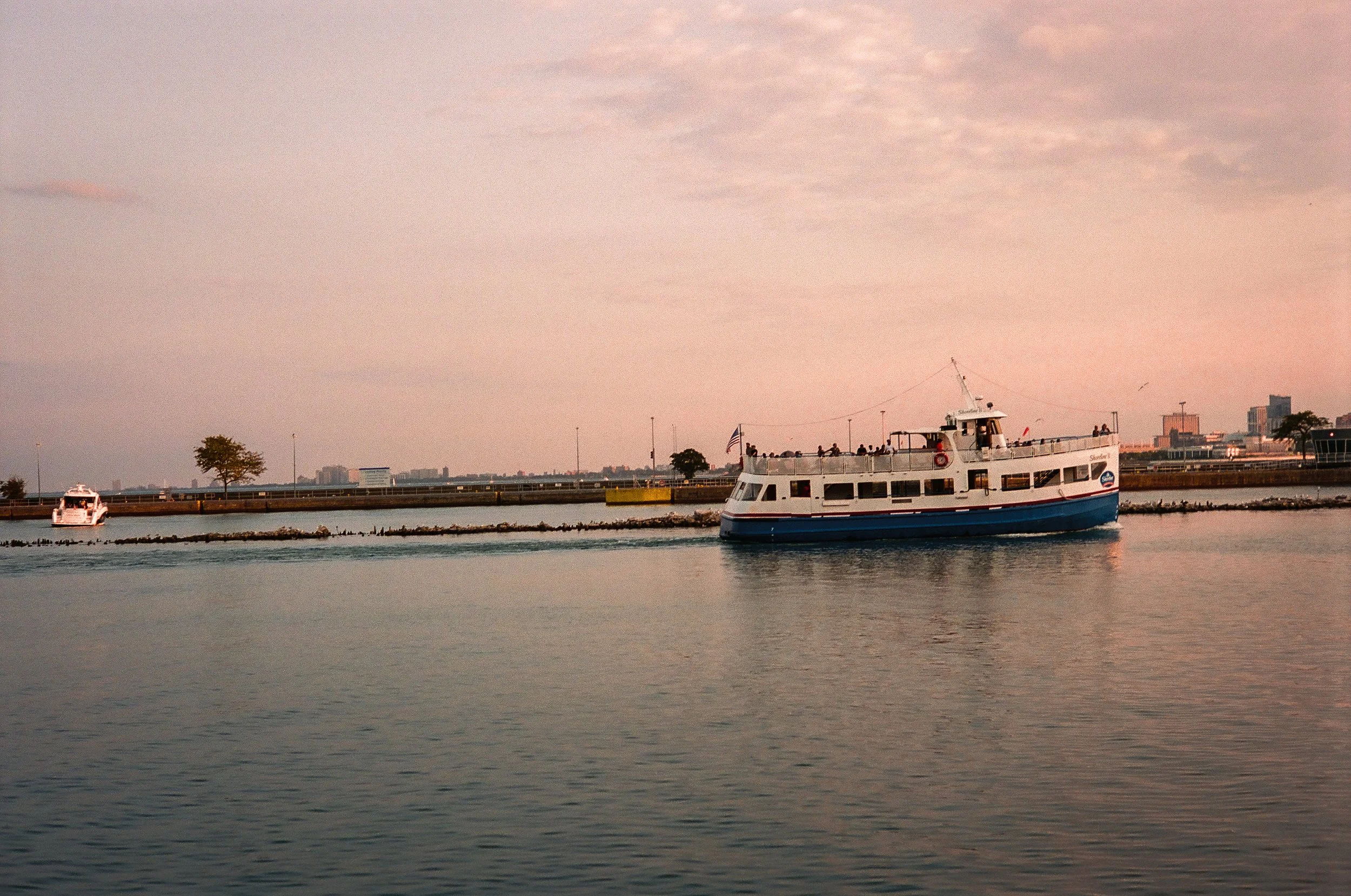 A boat sailing in a calm harbor at sunset with a city skyline in the background.