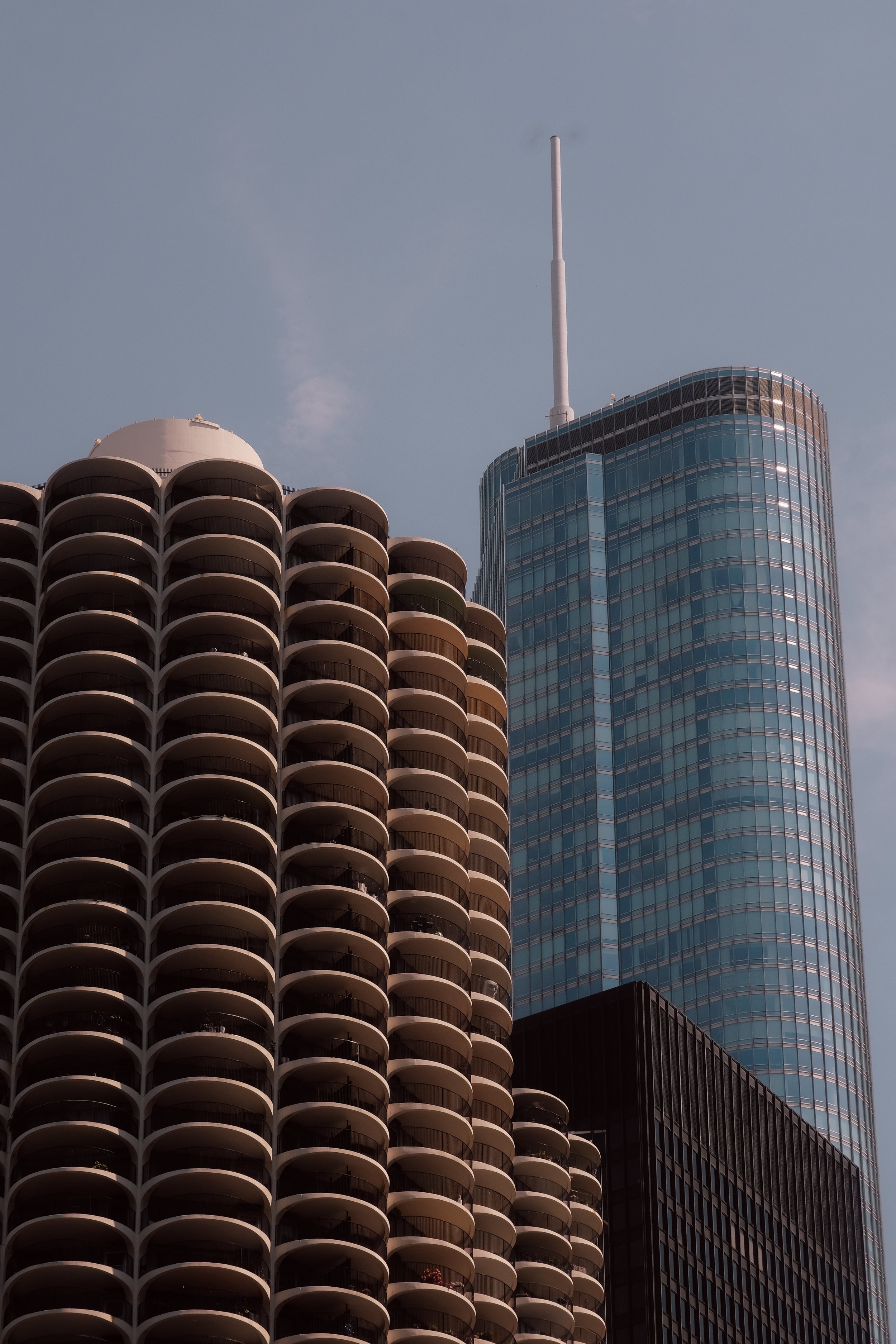 Close-up view of two modern skyscrapers with glass facades, one with curved balconies, and a taller building with a spire, set against a partly cloudy sky.