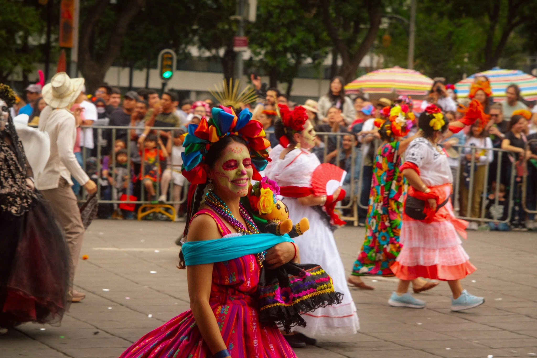 Woman with colorful traditional Mexican dress and Day of the Dead face paint holding a doll, walking in a parade with other women in festive costumes and face paint, crowd of onlookers watching behind barricades, some holding umbrellas.