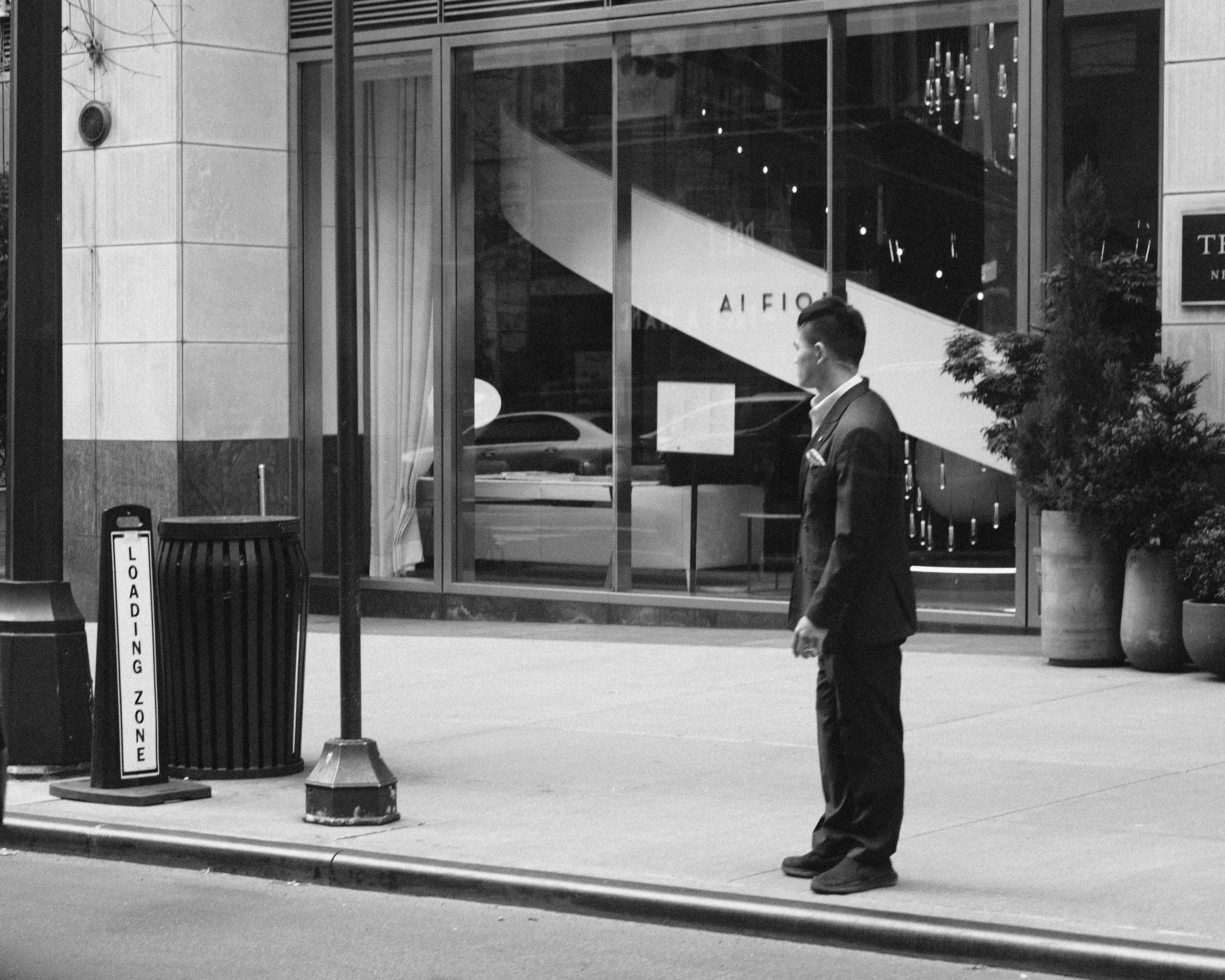 A man in a suit standing on a city sidewalk in front of a storefront with large glass windows, a potted plant, and a trash can labeled 'Loading Zone'.