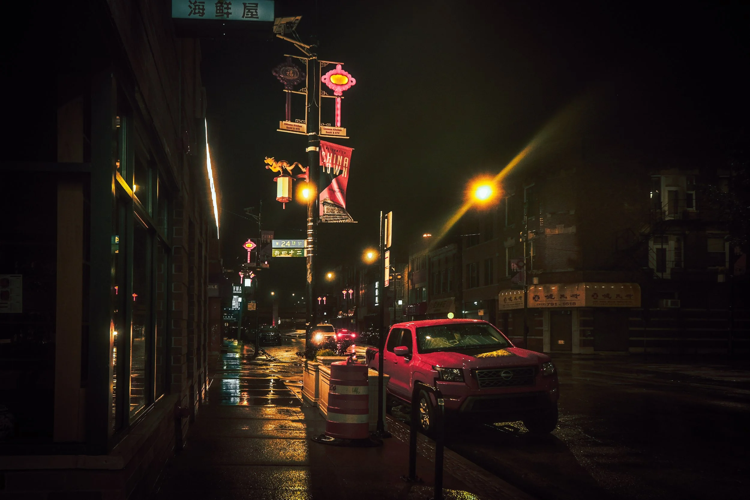 Night street scene with wet pavement, streetlights, and parked vehicles, including a pink Nissan truck. Decorative Chinese lanterns and banners hang from street poles, and signs indicate the area is Chinatown.