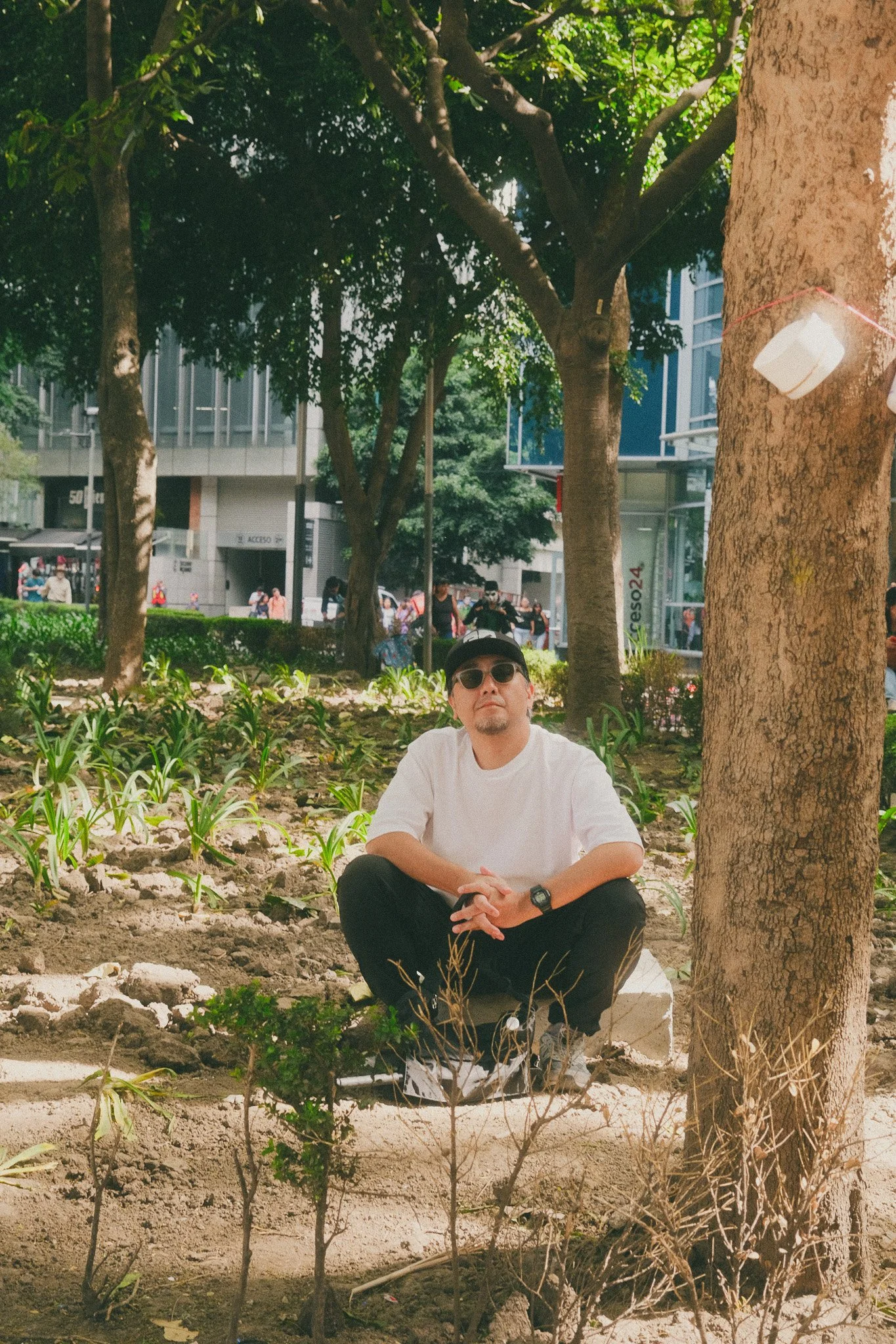 A man wearing sunglasses, a white T-shirt, and black pants, sitting on a small white stool among trees and plants in an urban park, with buildings and pedestrians visible in the background.