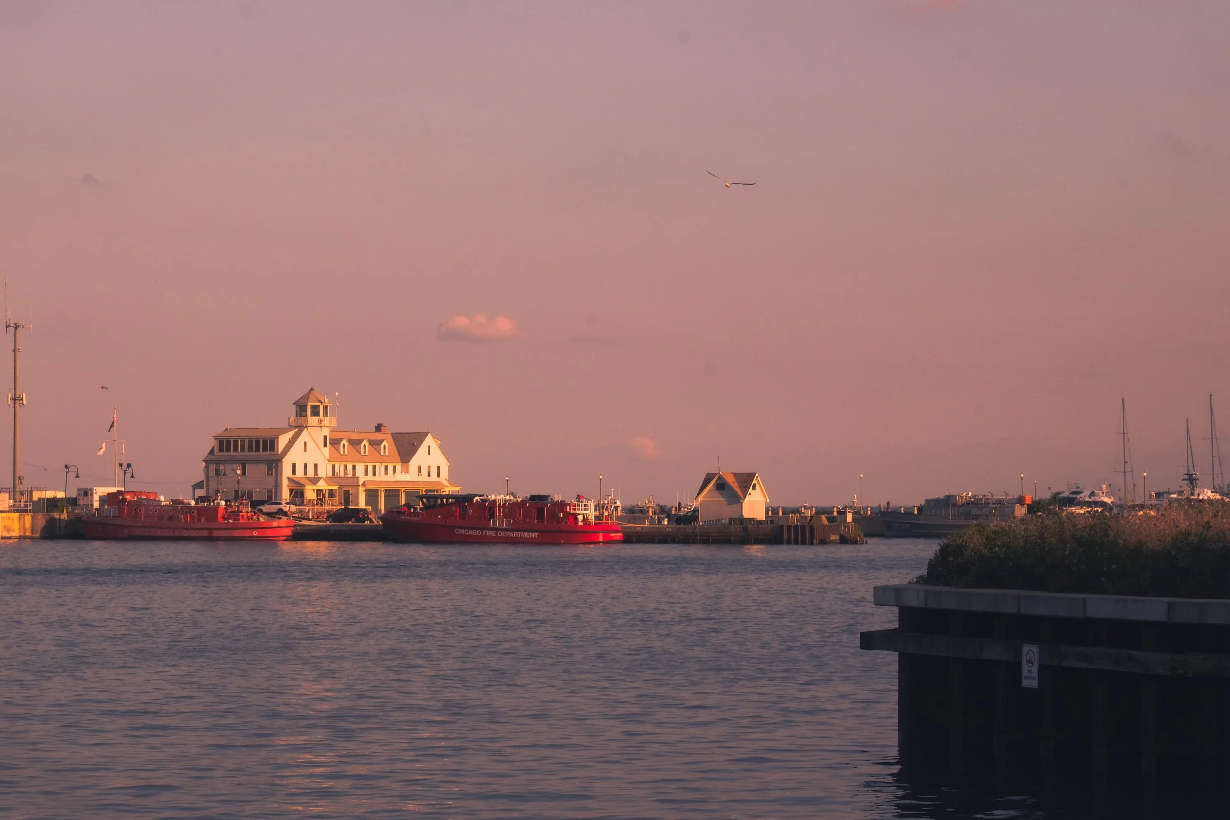 A waterfront scene at sunset with a large white building resembling a house or hotel, red fire boats docked in front, a small white building, and sailboats in the distance. A bird flies in the pinkish sky.