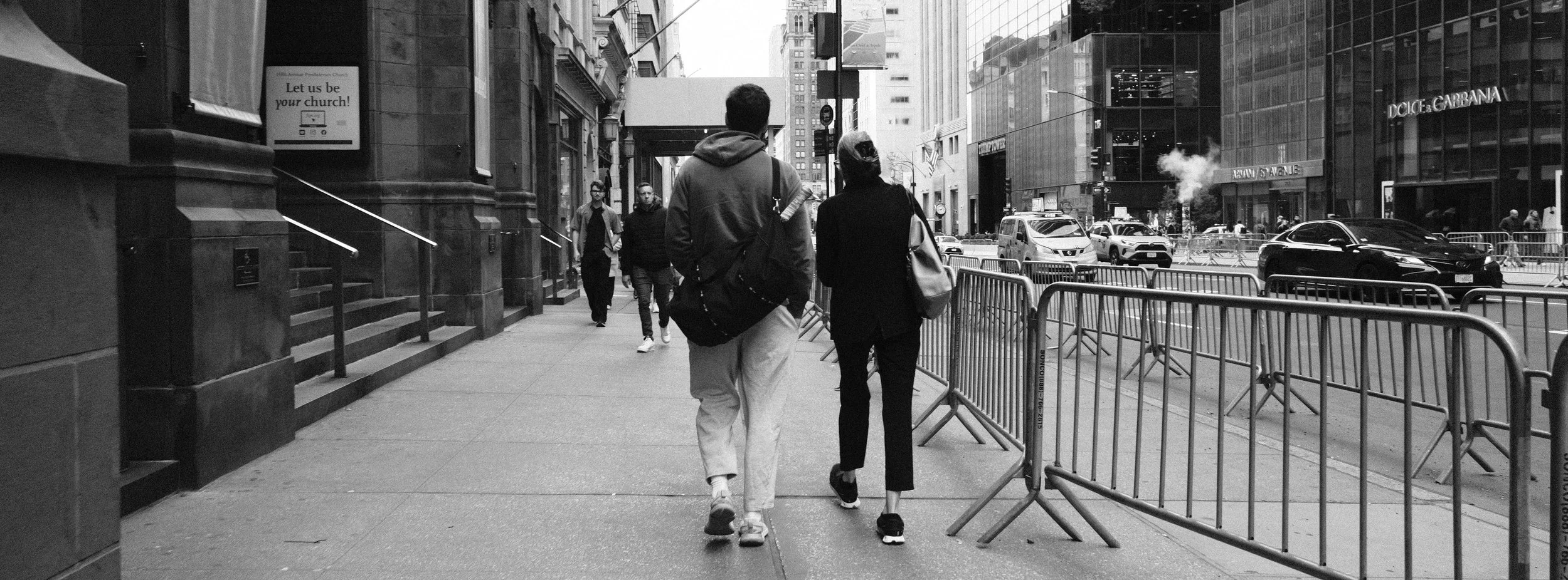 People walking on a city sidewalk with buildings and cars in the background, black and white photo.