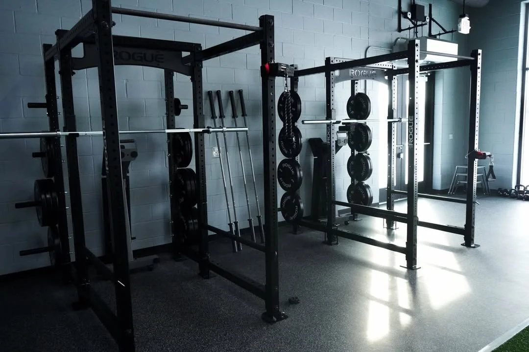 Image of a gym with weightlifting equipment including squat racks, barbells, and weight plates.