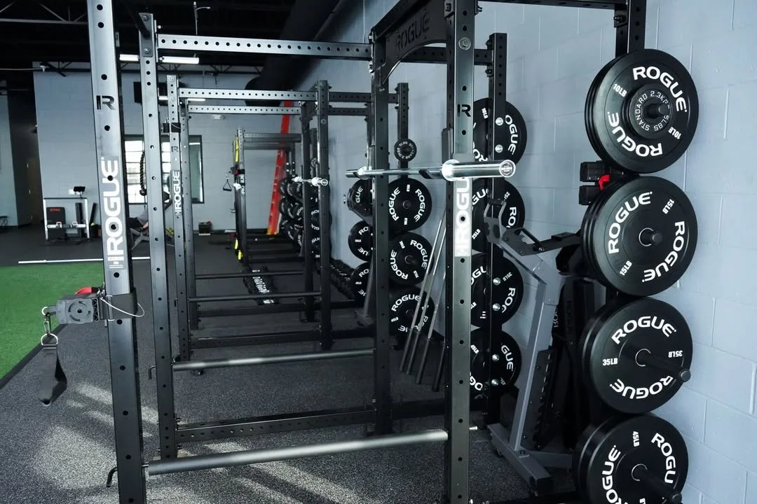 Inside a gym, a black power rack with weight plates and barbells. The wall to the right has stacked plates labeled 'Rogue' in black with white lettering. The floor has rubber mats, and in the background, there is a small green turf area.