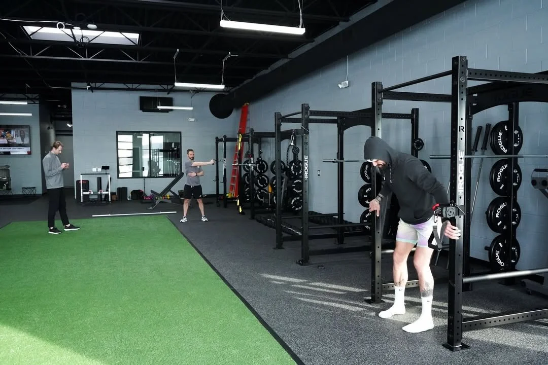 People working out in a gym with weightlifting equipment, an artificial turf area, and a man in a hoodie doing a workout.