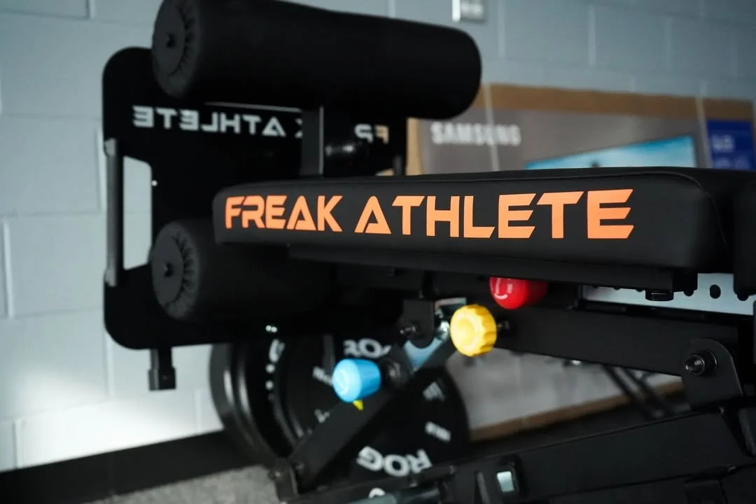 Close-up of a adjustable workout bench with the words 'FREAK ATHLETE' written on it, with weight plates and dumbbells in the background.
