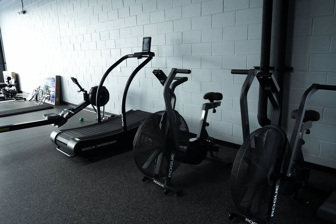 Gym area with a treadmill, stationary bike, and elliptical trainer, against a white brick wall.