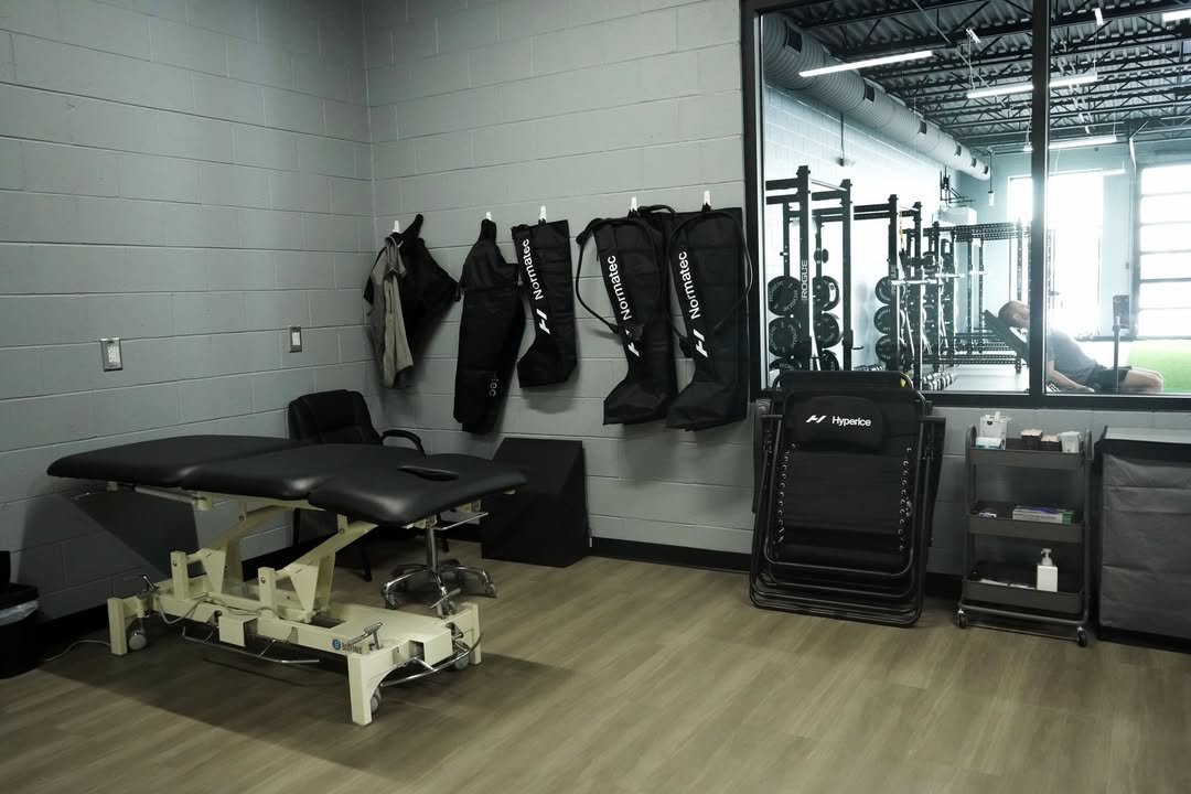 PT or therapy room with examination table, chairs, hanging bags, and gym equipment visible through a glass partition.