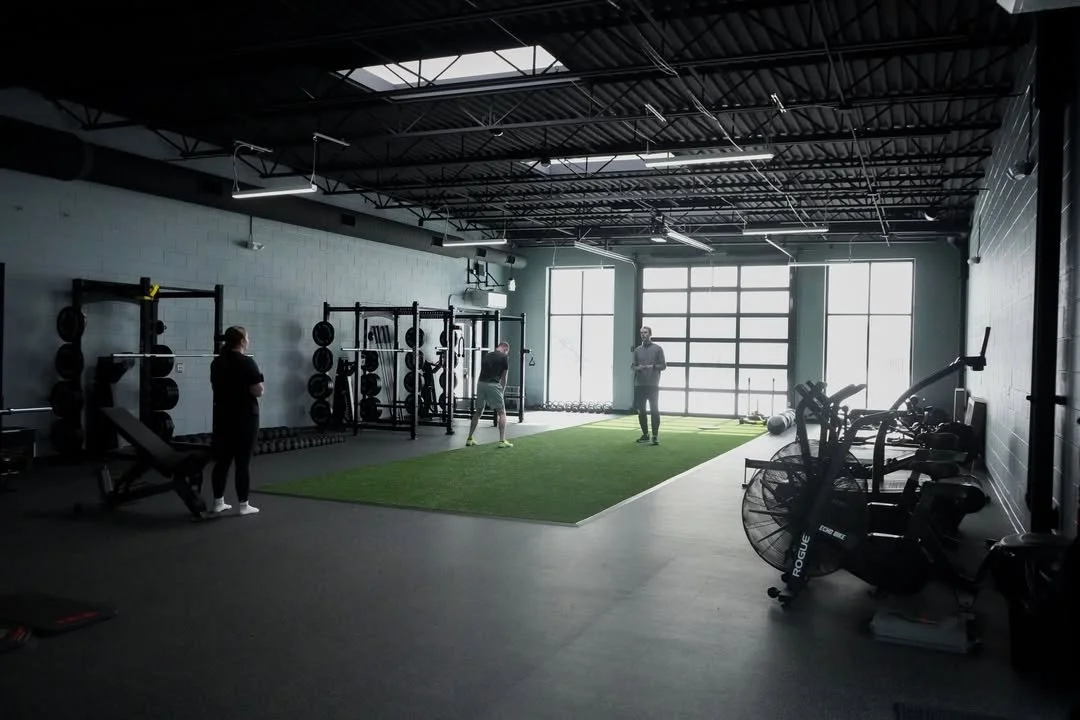 Interior of a modern gym with gym equipment, people exercising, and large windows at the back.
