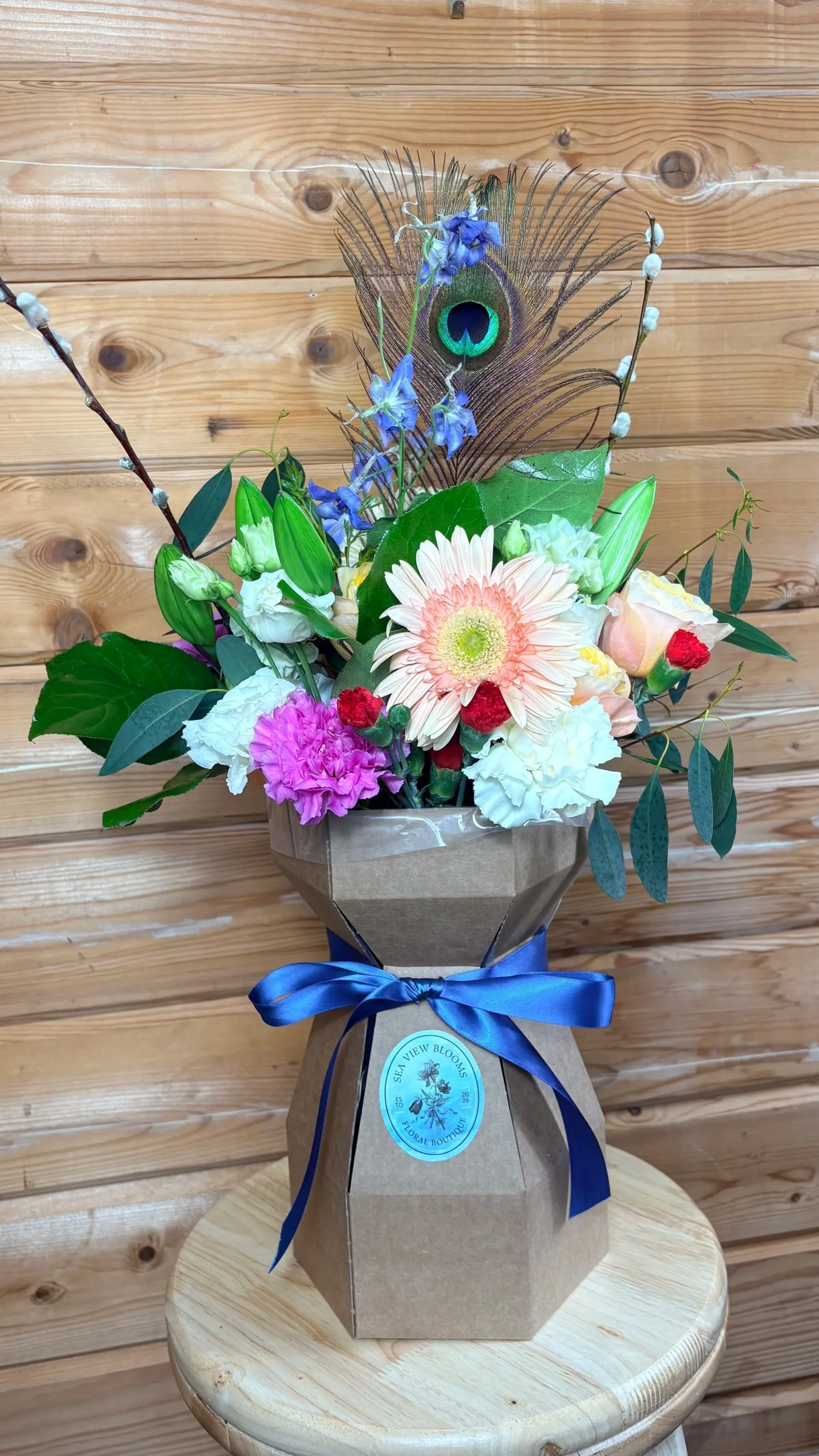A flower arrangement with peacock feathers, blue and purple flowers, and green leaves in a brown gift bag tied with a blue ribbon, placed on a small wooden table against a wooden wall background.