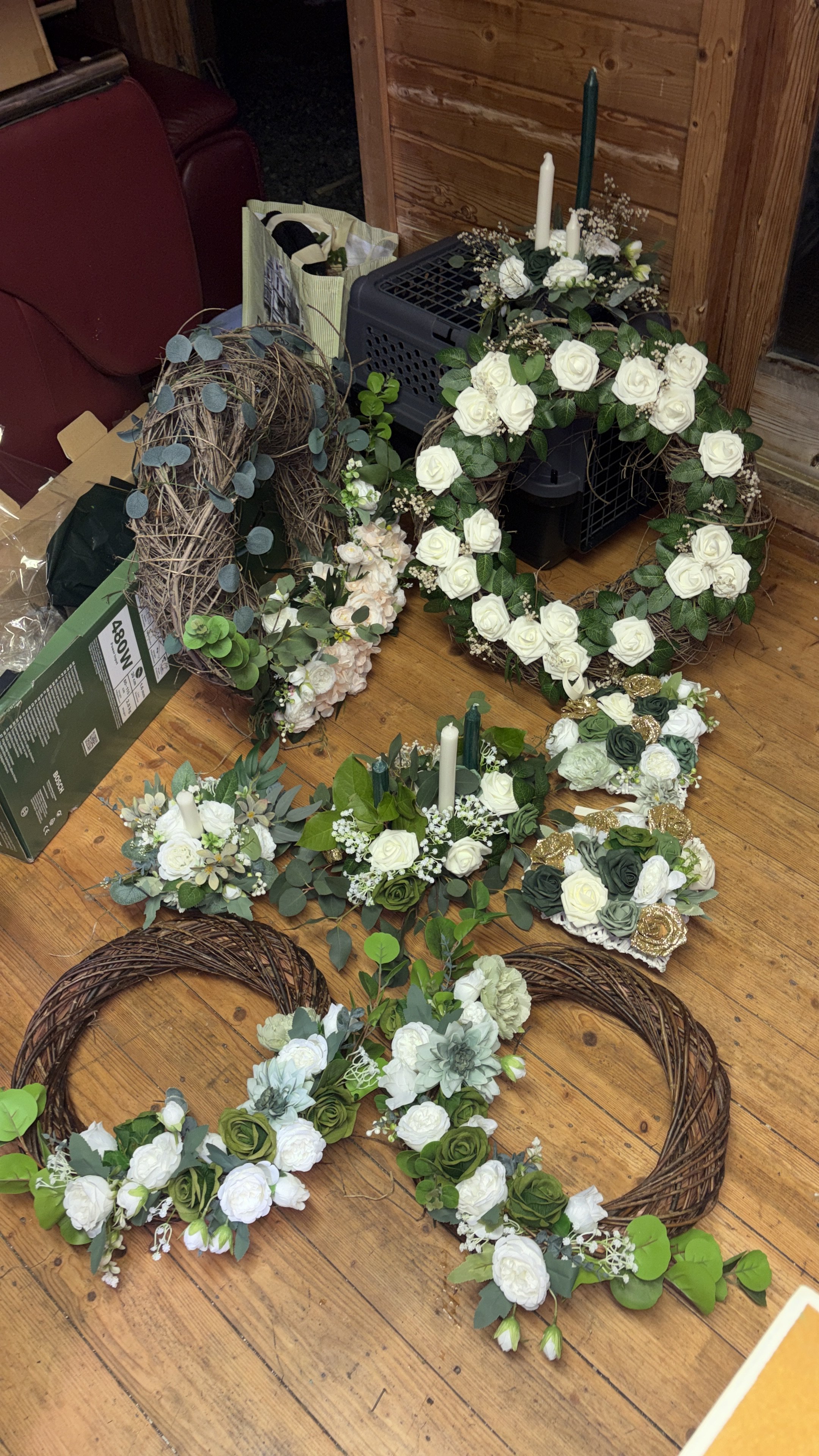 Various decorative floral wreaths and arrangements with white, green, and cream-colored flowers and greenery on a wooden floor, including some grapevine wreaths and candle decorations.