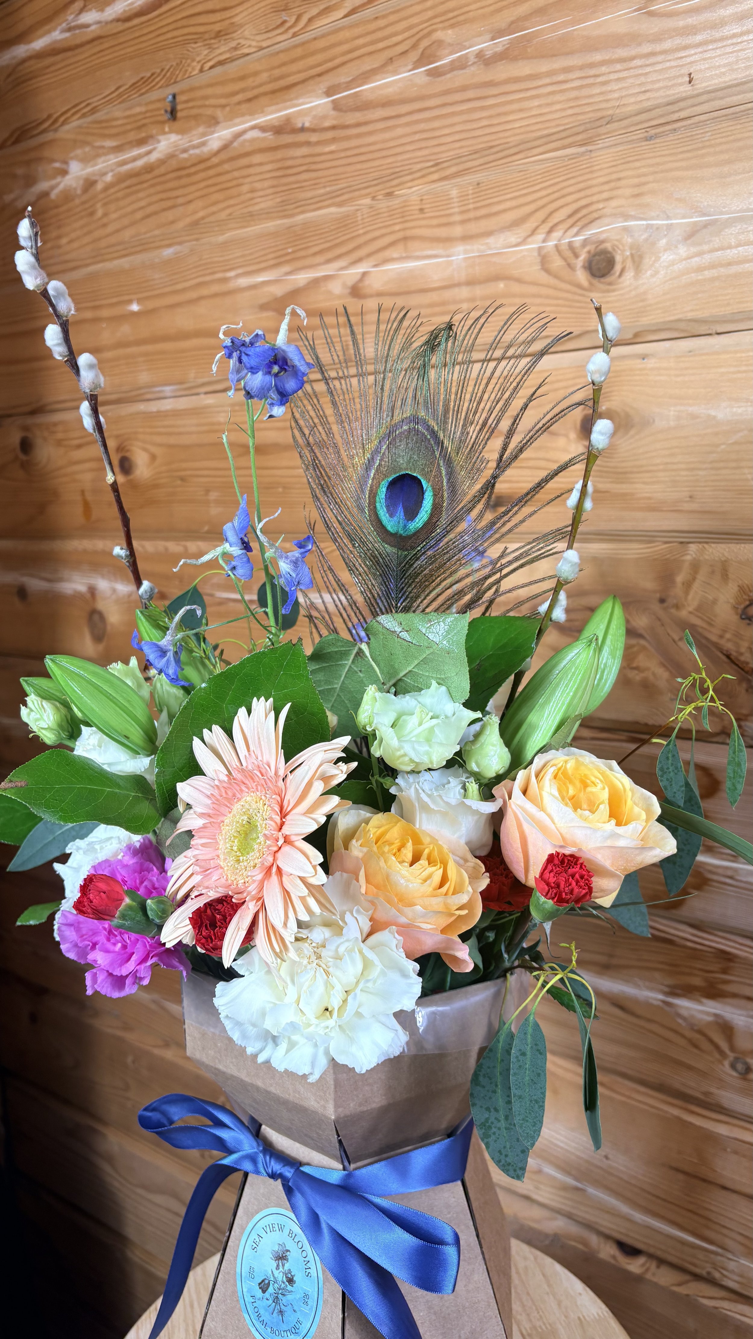 A bouquet of mixed flowers including peach roses, pink carnations, white lisianthus, gerbera daisy, blue delphinium, and a peacock feather, wrapped in craft paper with a blue ribbon.