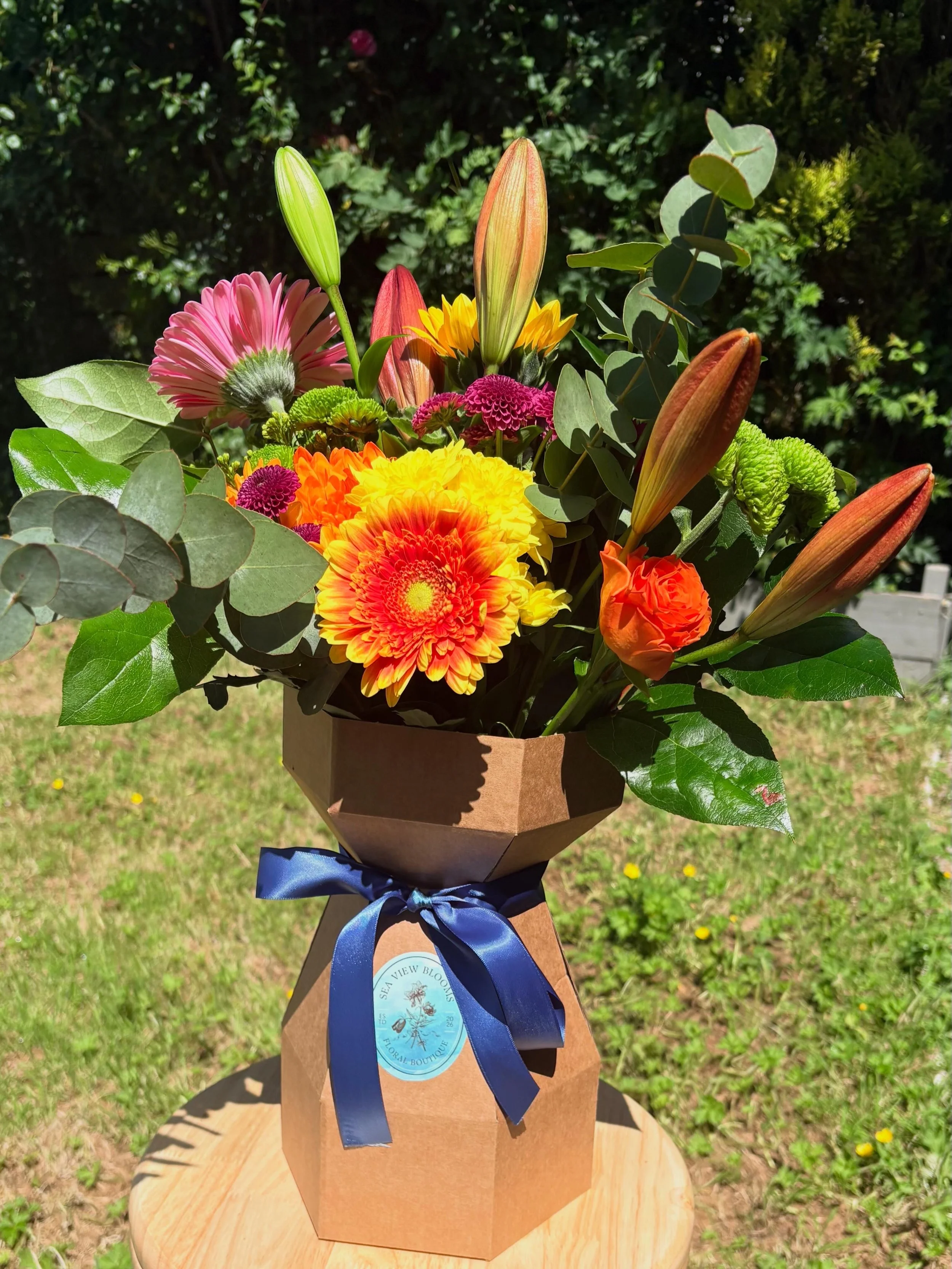 A colorful bouquet of fresh flowers, including lilies, daisies, and other blooms, wrapped in brown paper with a blue ribbon and a round blue tag. The bouquet is outdoors on a wooden surface with green grass and plants in the background.