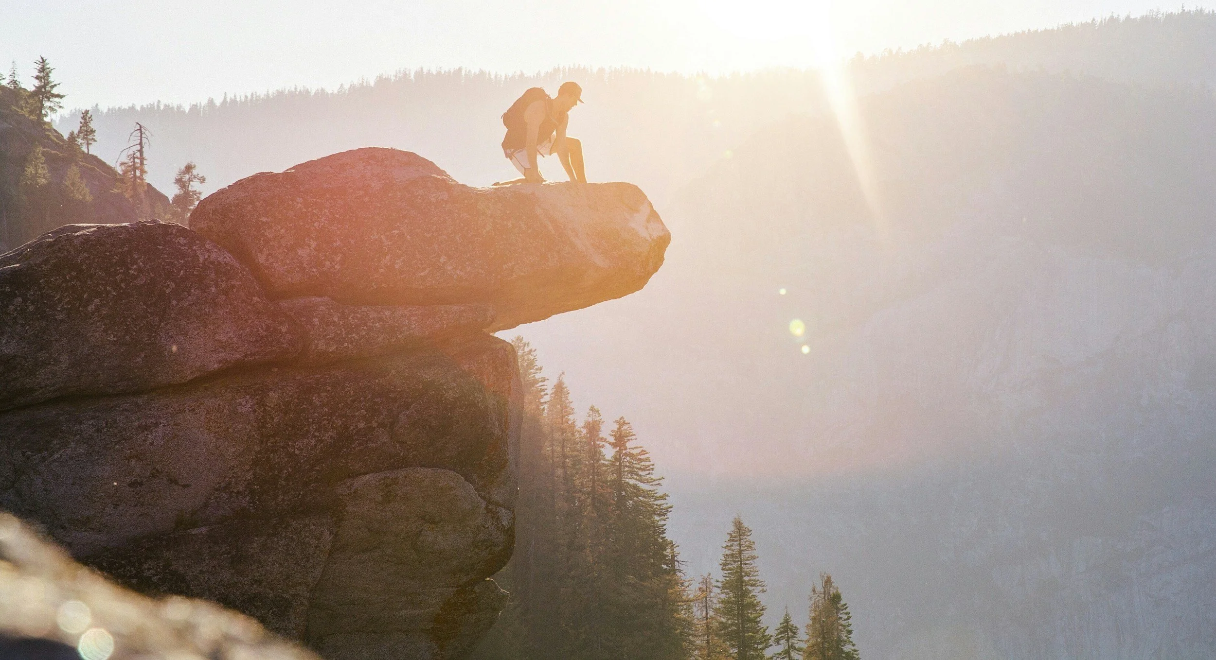 A person with a backpack is crouching on the edge of a large rock formation overlooking a mountain landscape during sunset.
