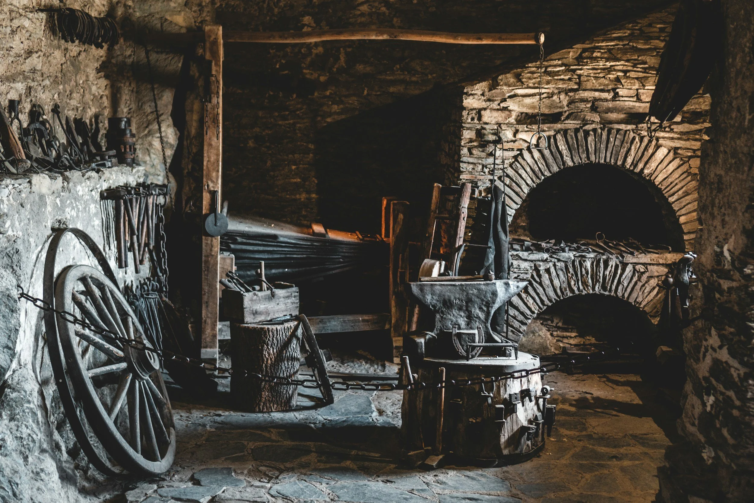 A rustic blacksmith's workshop with antique tools, an anvil, and a stone oven. The scene is dimly lit, emphasizing the historic and industrial atmosphere.