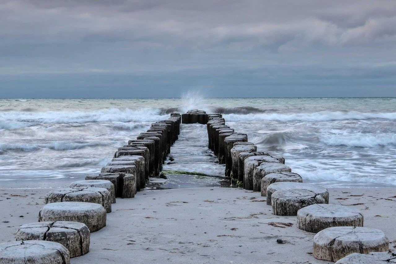 Wooden breakwater extending into the ocean with waves crashing against it under a cloudy sky.