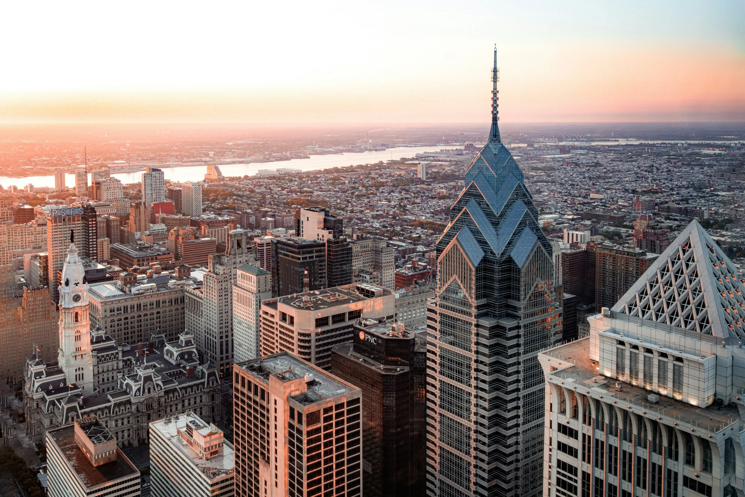 Aerial view of downtown Philadelphia at sunset with tall skyscrapers, including the Liberty Place buildings, and the Schuylkill River in the background.