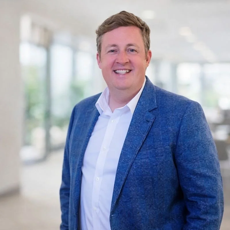 A man smiling in a professional setting, wearing a blue blazer and white shirt, standing in a bright, modern office.
