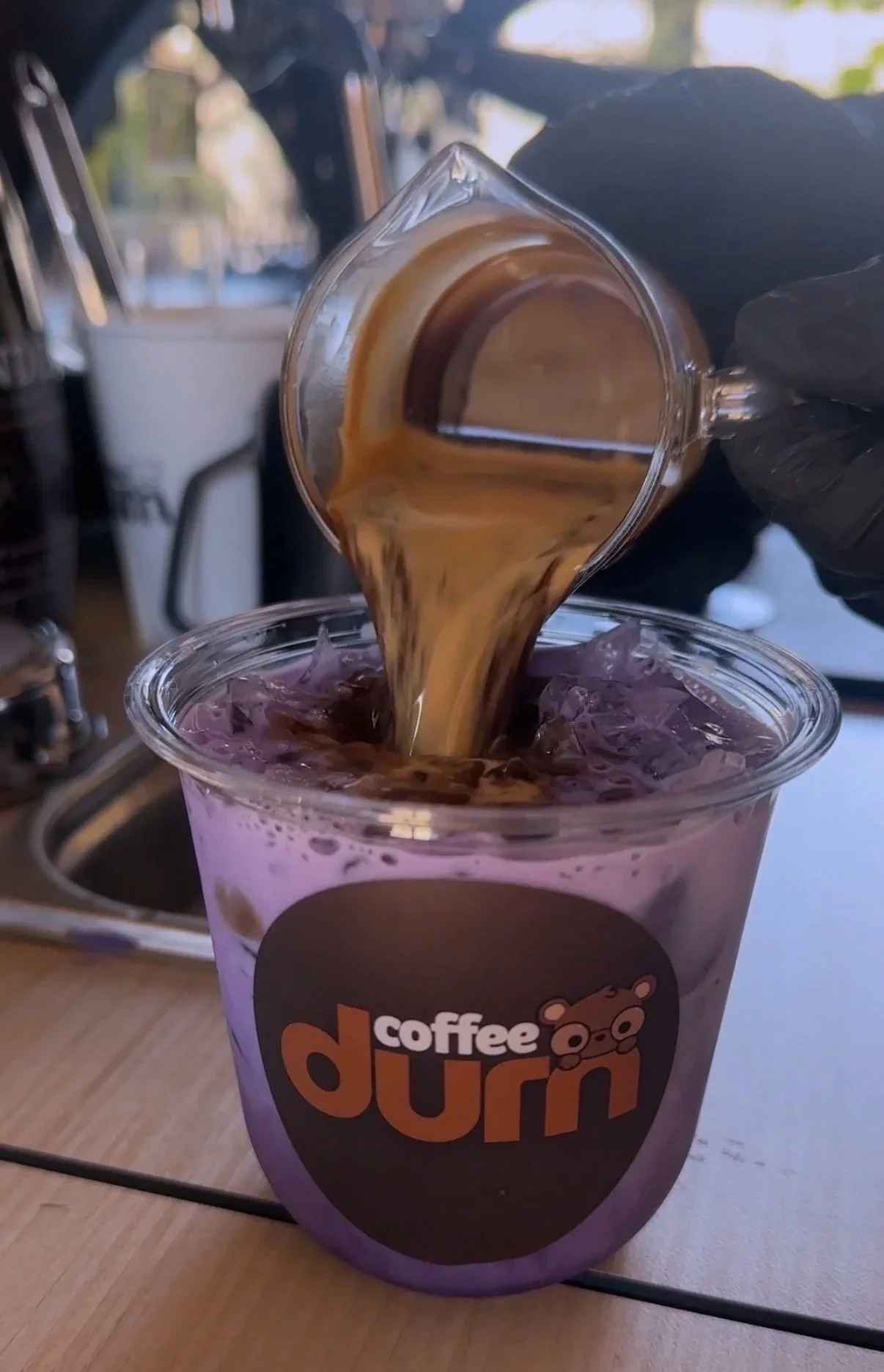 Coffee drink with ice being poured into a purple cup labeled 'coffee durr', on a wooden surface, with a blurred background of a café setting.