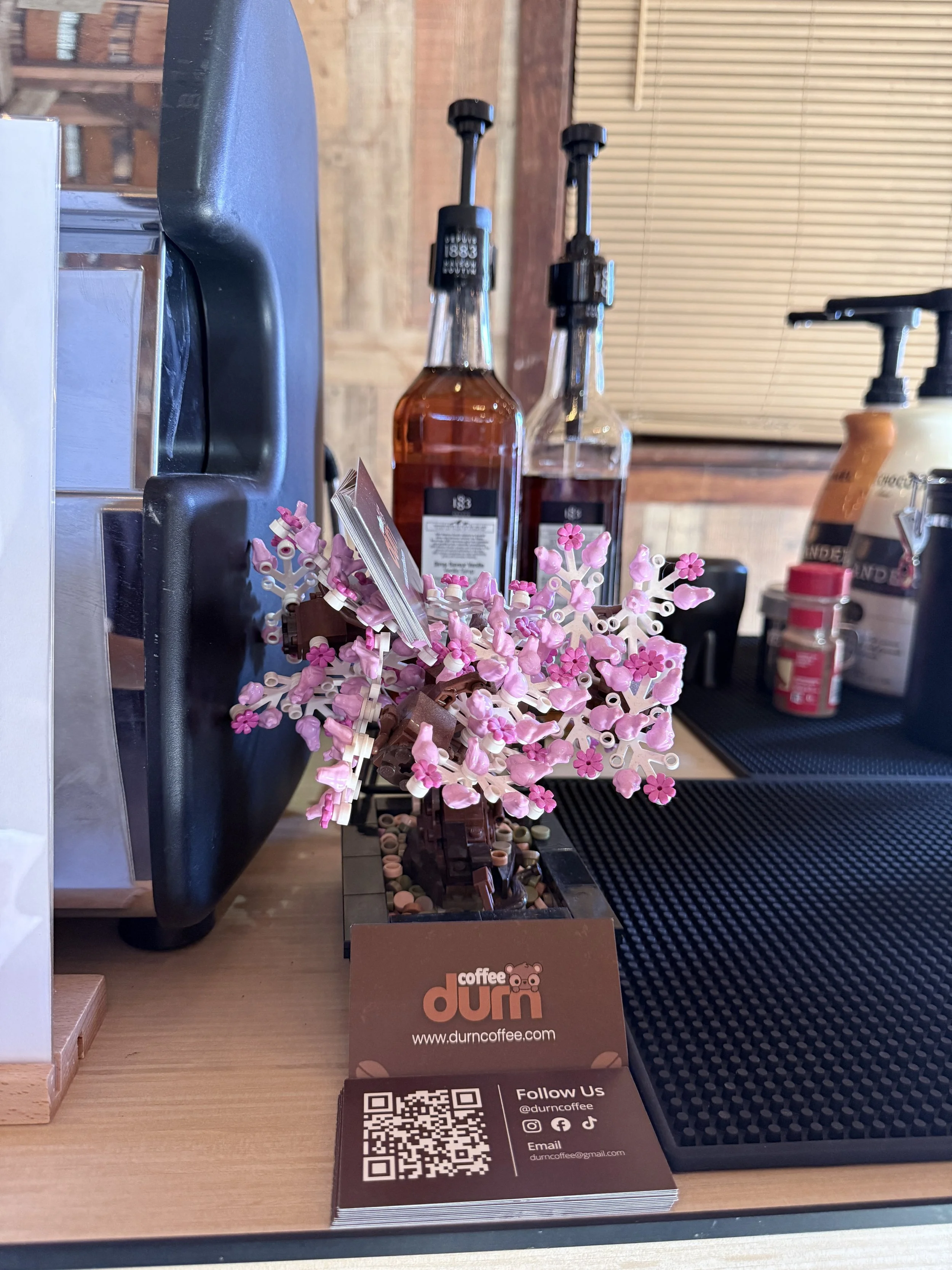 Business card for DÙRN Coffee with logo of a bear, QR code, social media icons, and email address on a wooden surface. In the background, there are bottles of syrup, a small black plastic container, and a decorative pink and white tree-like object ma