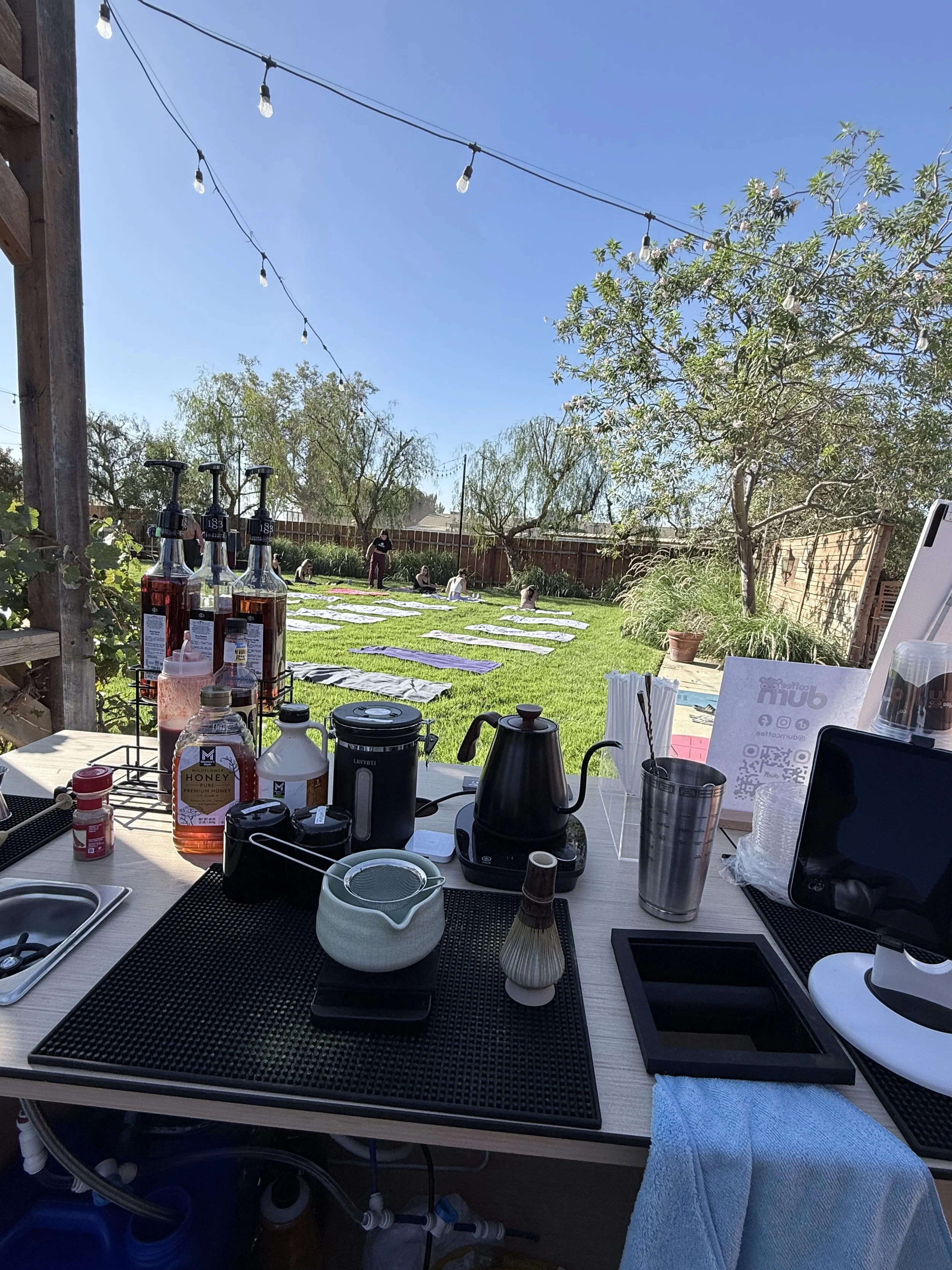 Outdoor garden scene with yoga mats and people practicing yoga, string lights overhead, trees and a wooden fence, and a table with various drinks and equipment in the foreground.