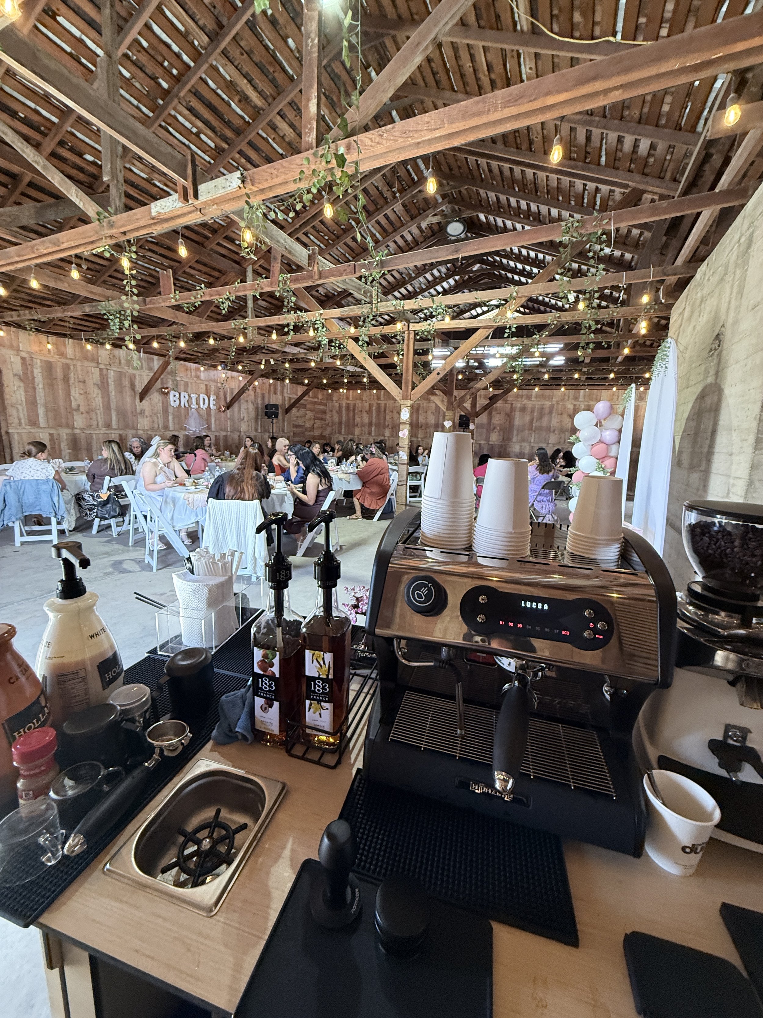 Inside a rustic wedding reception barn with wooden beams and string lights, decorated with balloons and a 'BRIDE' sign, people socializing at tables, coffee station with cups, coffee machine, and syrup bottles in the foreground.