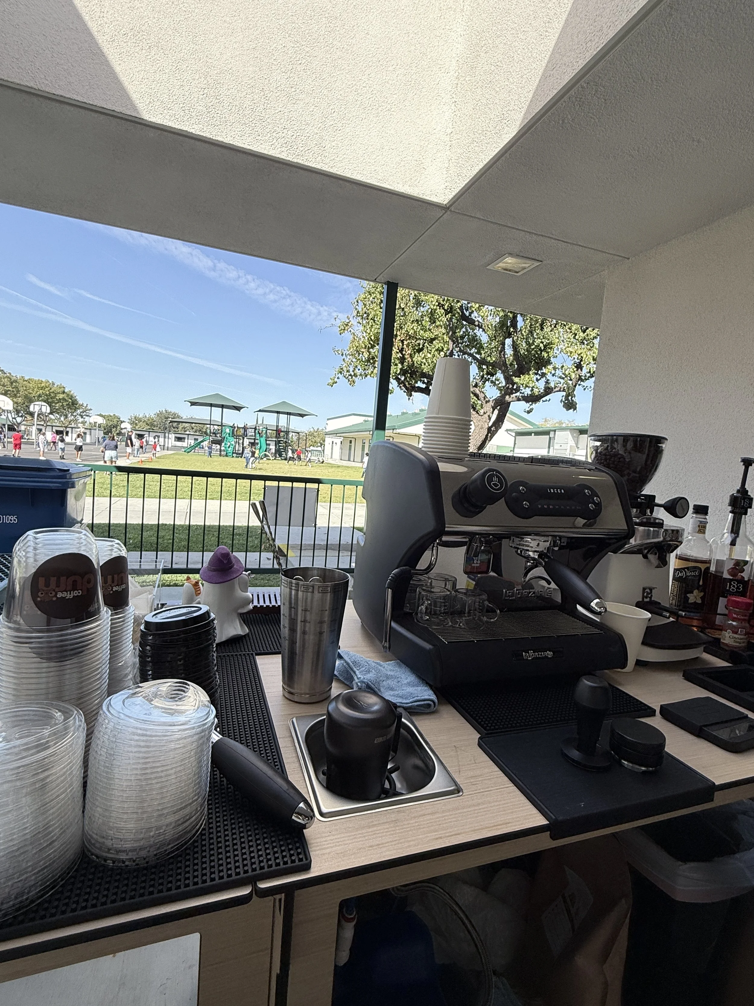 A coffee stand on a patio with a barista espresso machine, stacked cups, and various supplies, overlooking a playground and children playing outside.