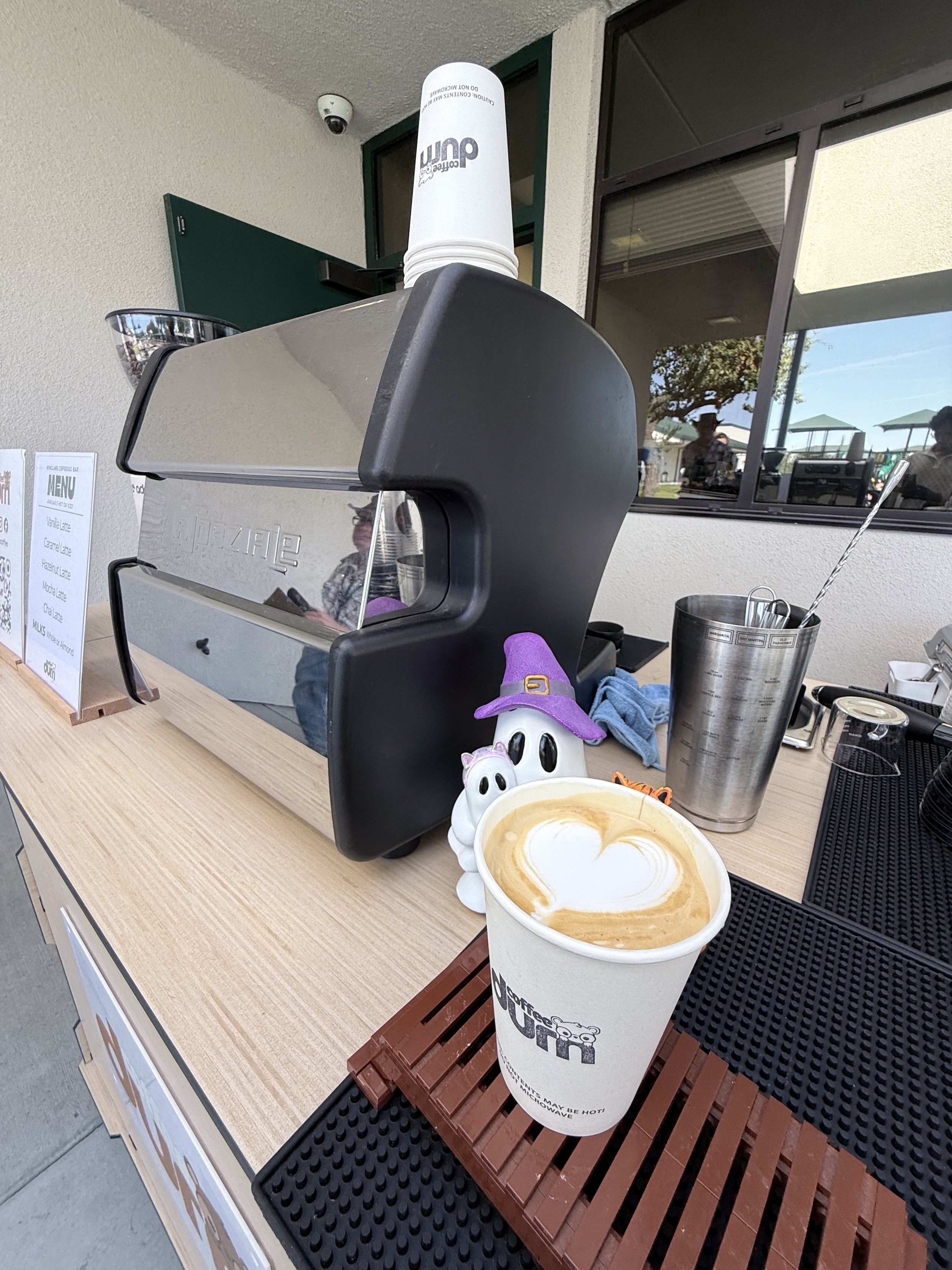A coffee cup with latte art resting on a countertop in front of a coffee machine. There is a small witch hat decoration on the cup. The coffee shop window reflects outdoor seating and people.