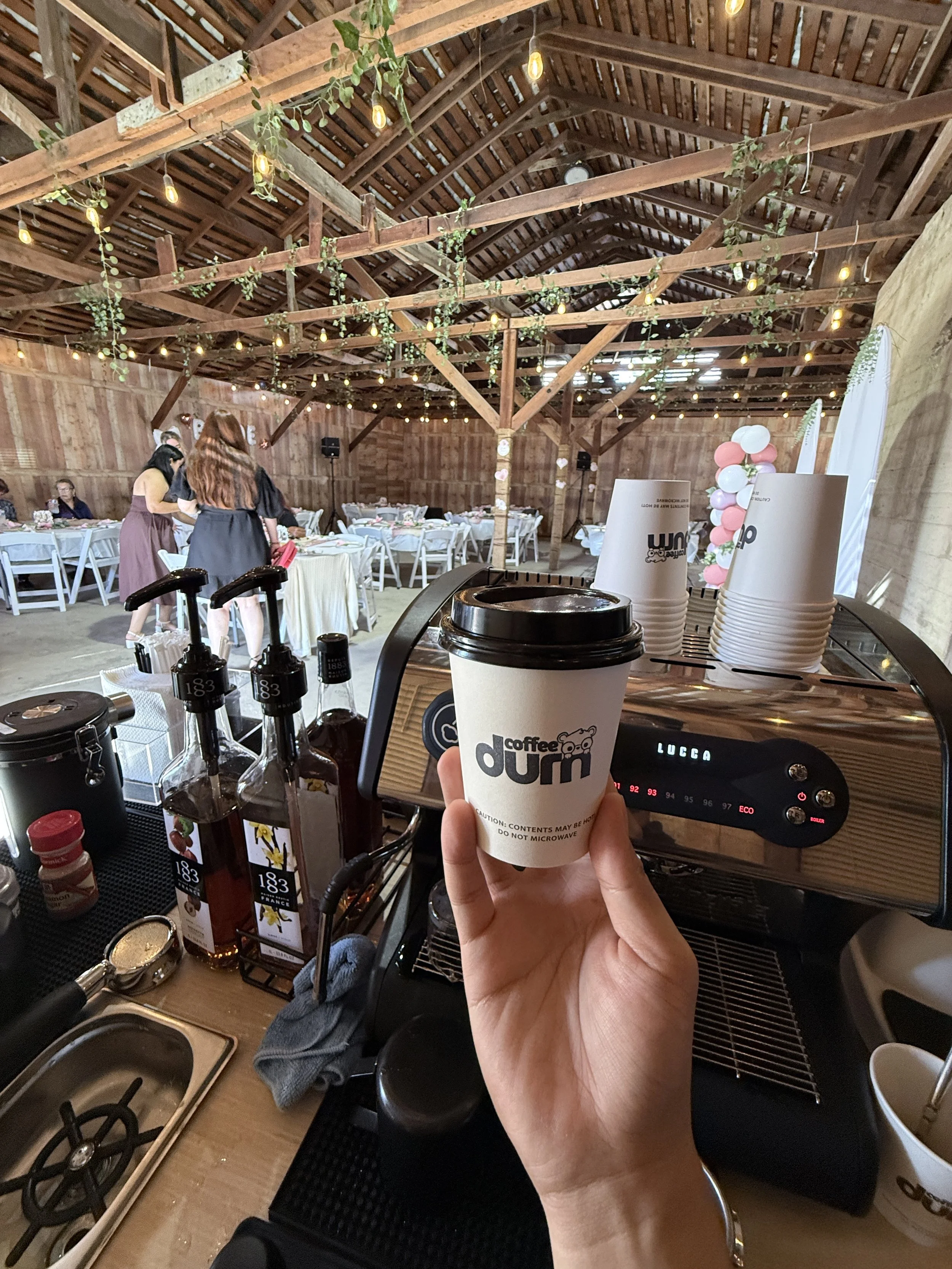 A person holding a coffee cup in front of a coffee machine, with a rustic wooden event space in the background decorated with string lights and balloons.