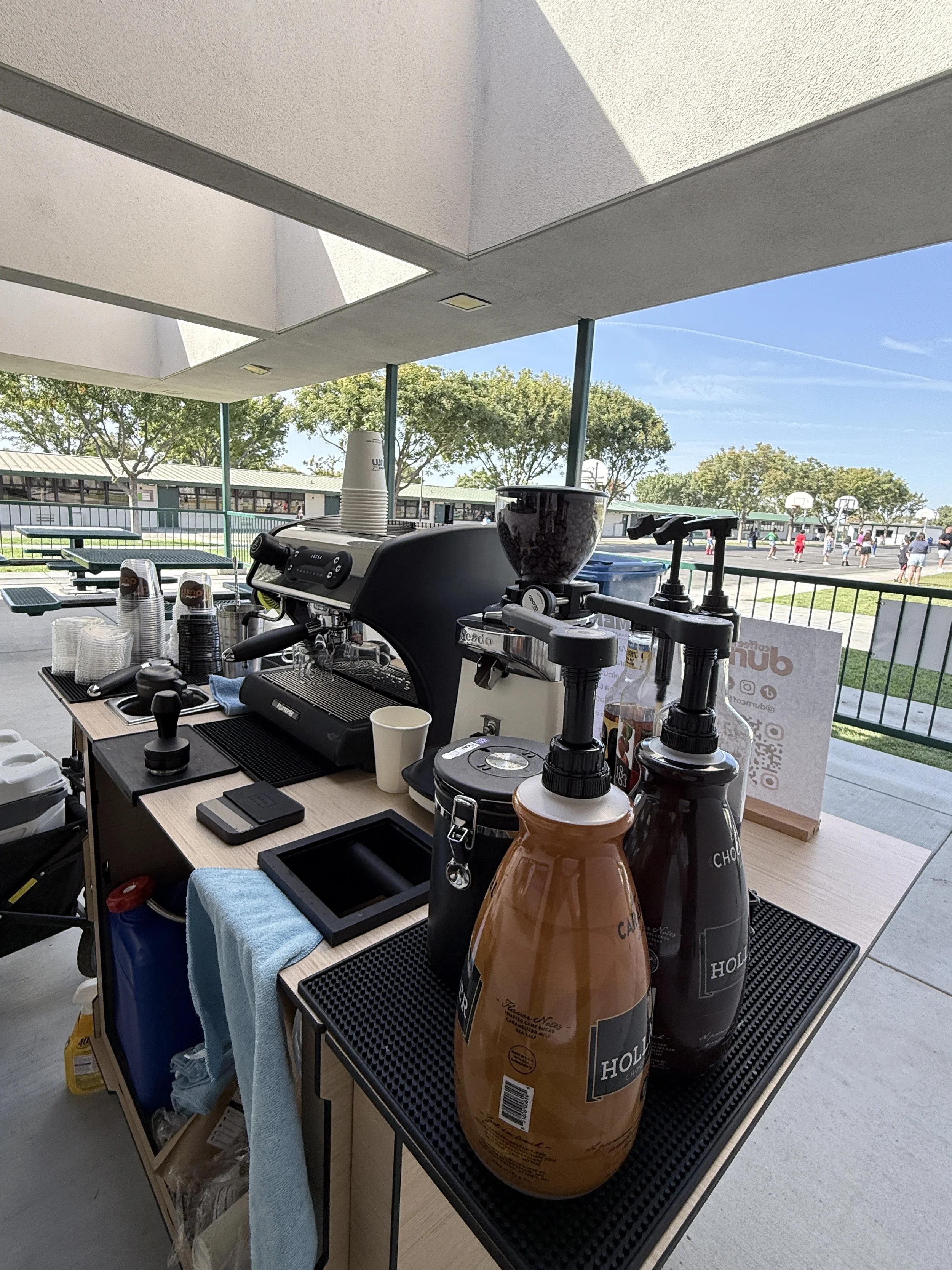 A coffee station set up outdoors with coffee equipment, cups, and syrup bottles, overlooking a basketball court and trees under a clear blue sky.