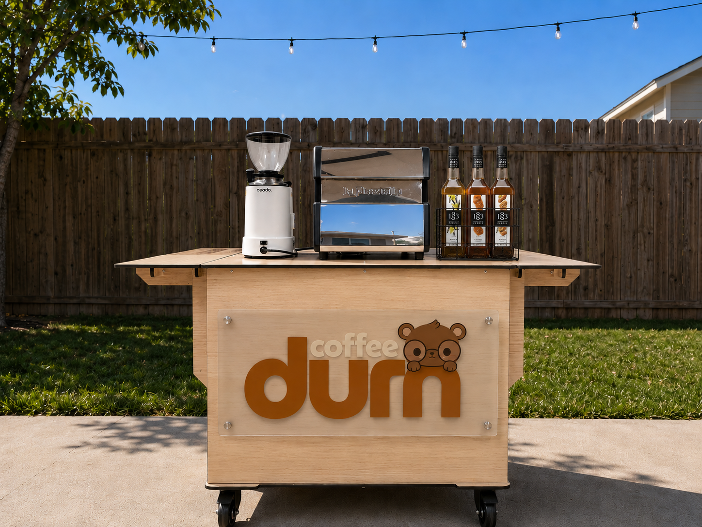 A mobile coffee stand with a wooden front panel displaying a bear logo and the words 'coffee durm'. On top, there is a white coffee grinder, a silver toaster, and three bottles of coffee syrup. String lights are hanging above, and a wooden fence with a tree and blue sky is in the background.