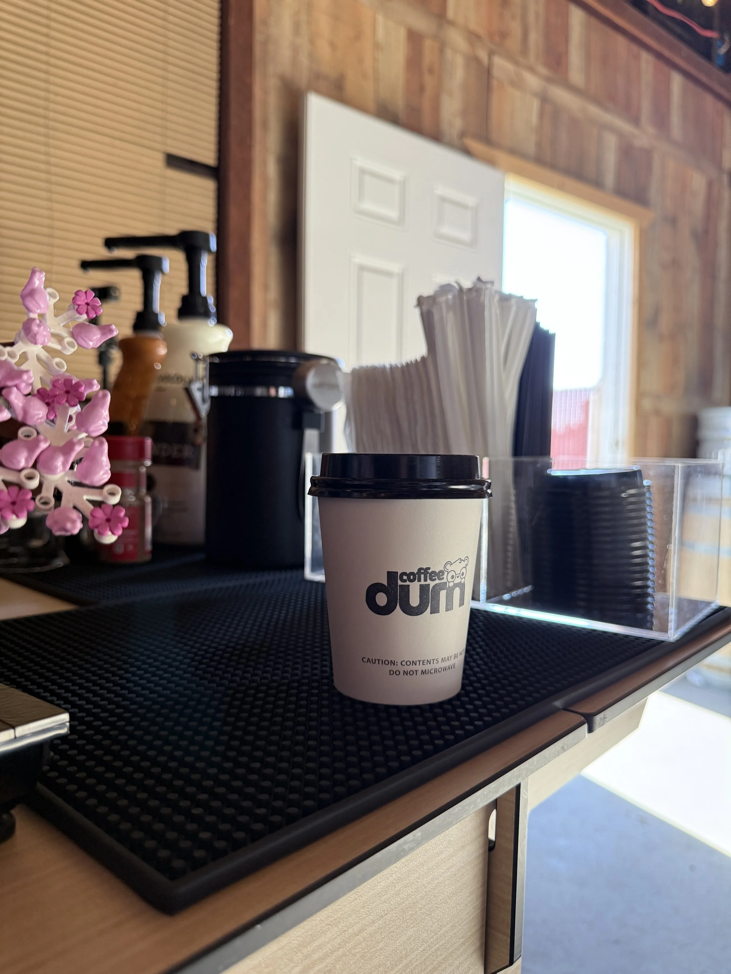 A paper coffee cup with a black lid on a black rubber mat, with pink and white decorative flowers and soap dispensers nearby on the counter. There is a window and wooden wall in the background.