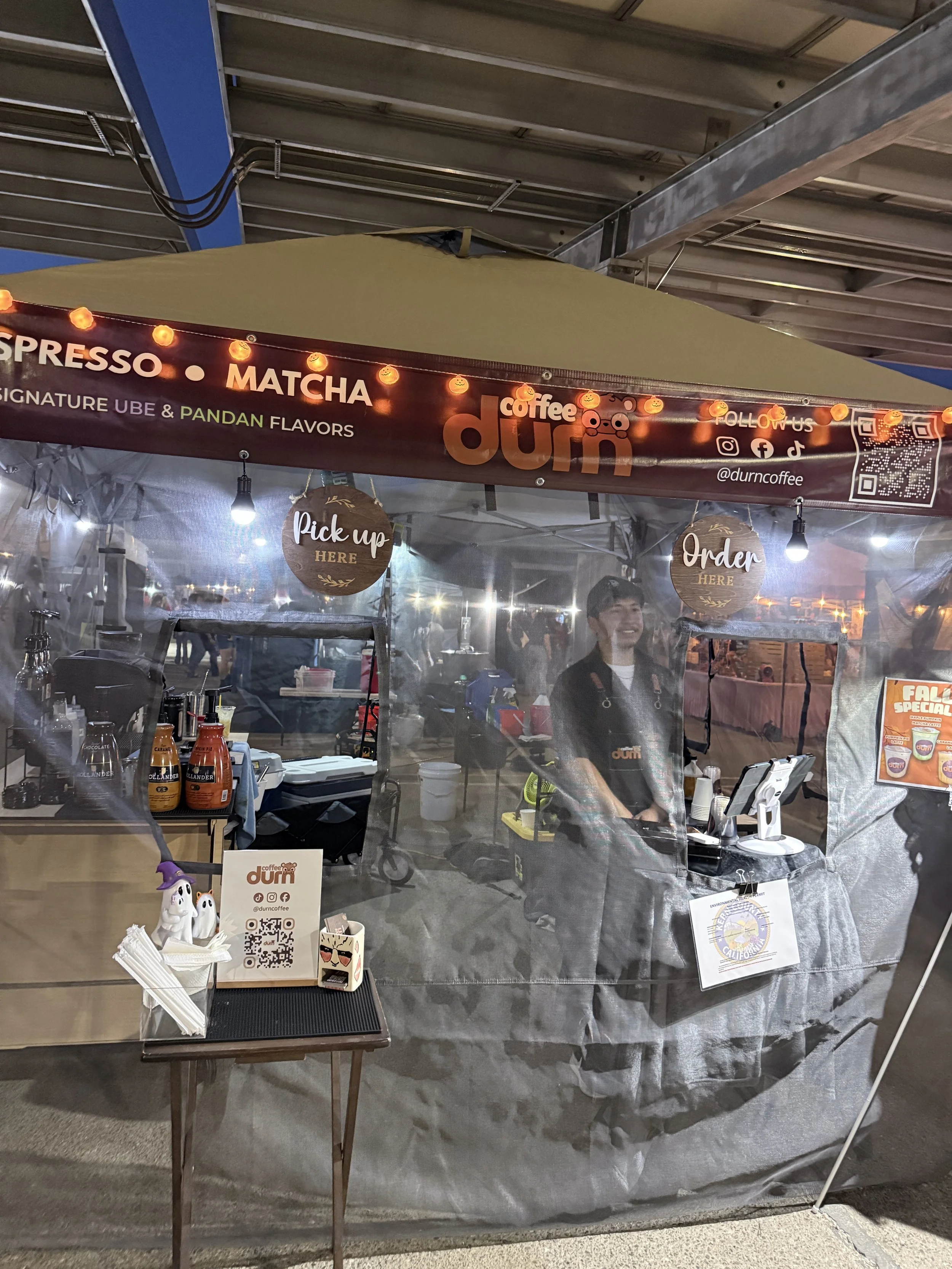 A food stand at an outdoor market with signs for coffee, matcha, and espresso, decorated with small pumpkin lights, and staff ready to serve customers.
